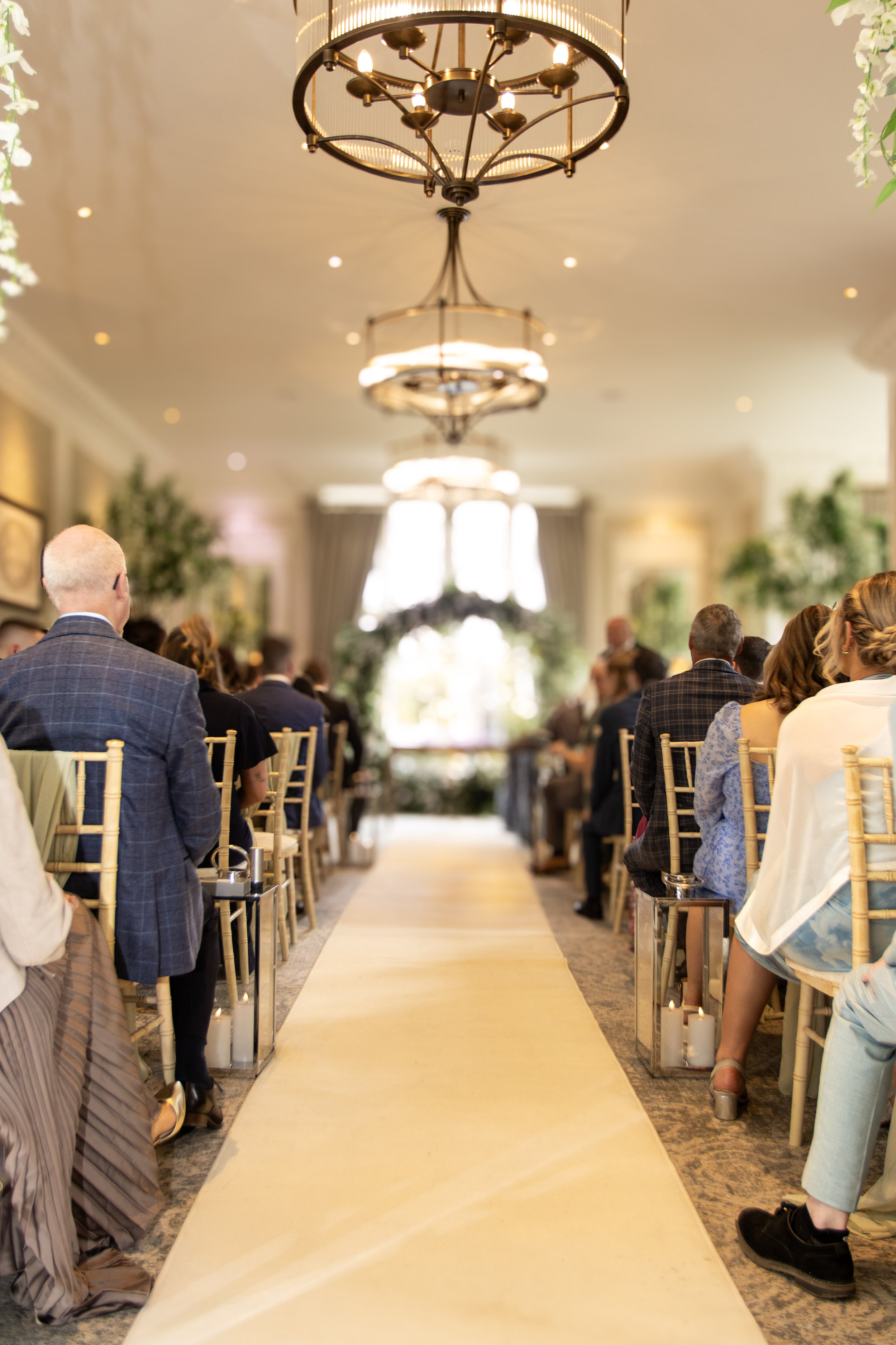 A wedding ceremony in a beautifully decorated indoor venue with a yellow aisle runner, surrounded by guests seated on gold chairs, chandeliers hanging from the ceiling, and natural light coming from large windows at the end of the aisle.