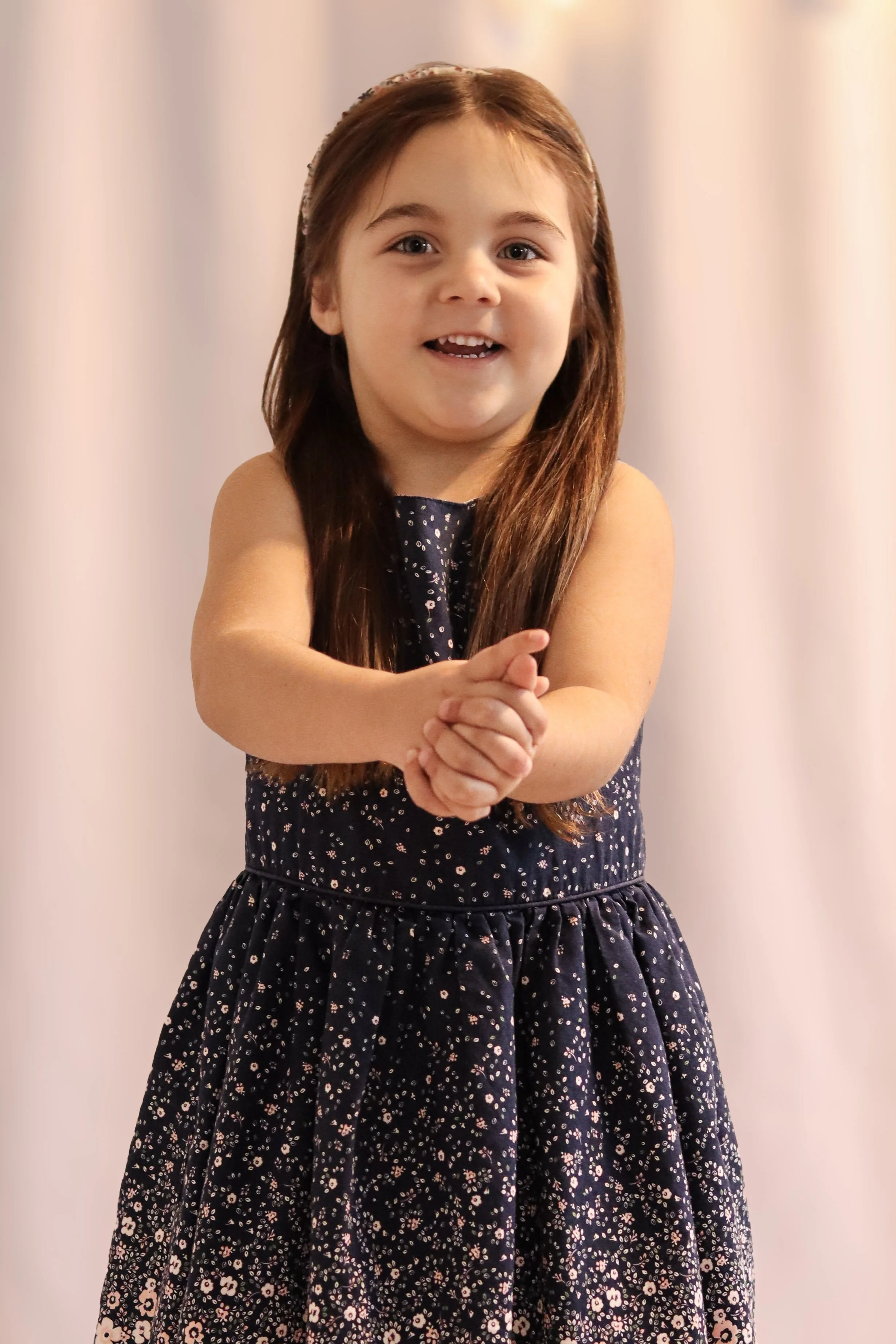 A young girl with long brown hair, smiling, wearing a navy blue dress with small floral print, standing with her hands clasped together in front of her.