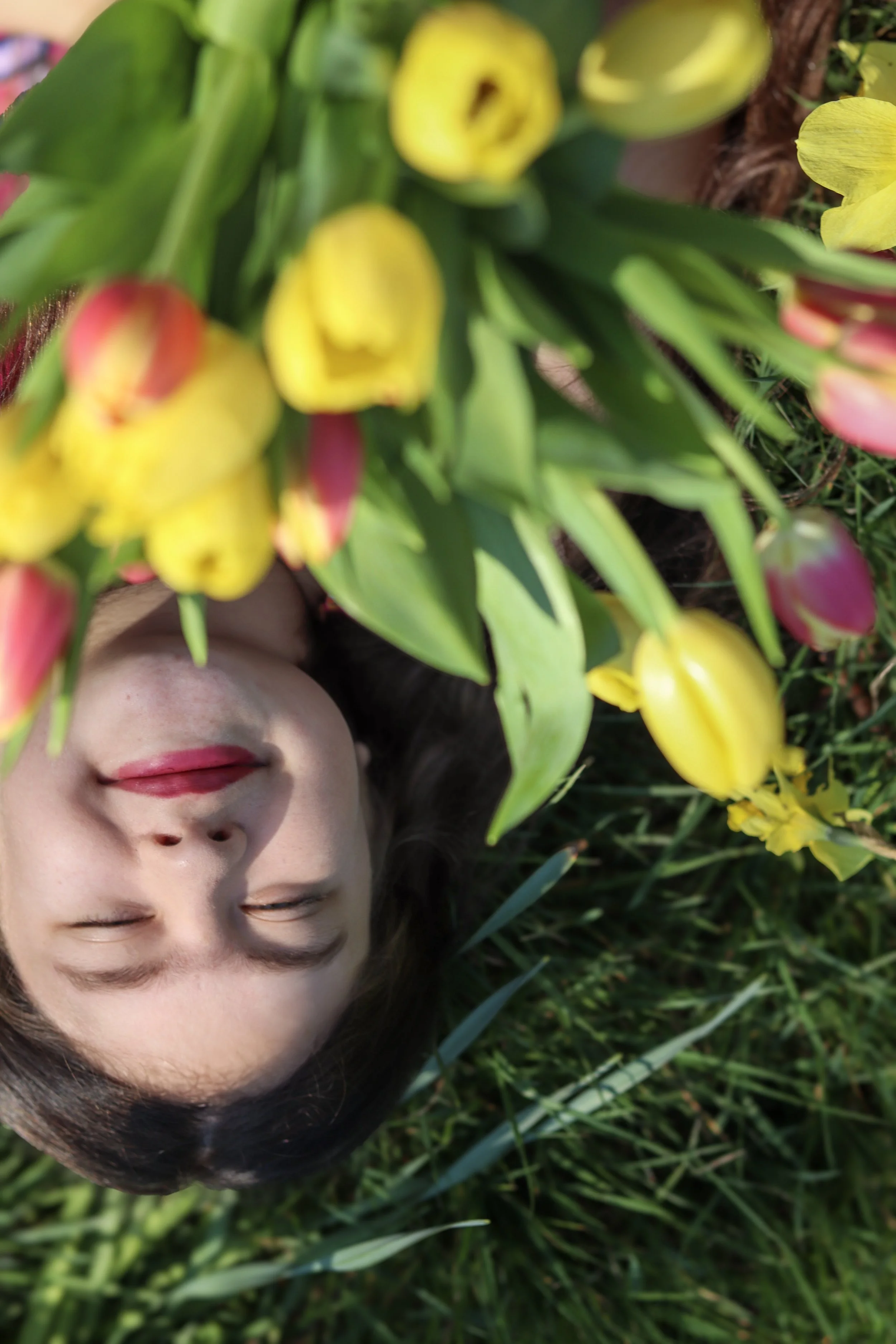 A woman lying on grass with eyes closed, surrounded by yellow and pink flowers