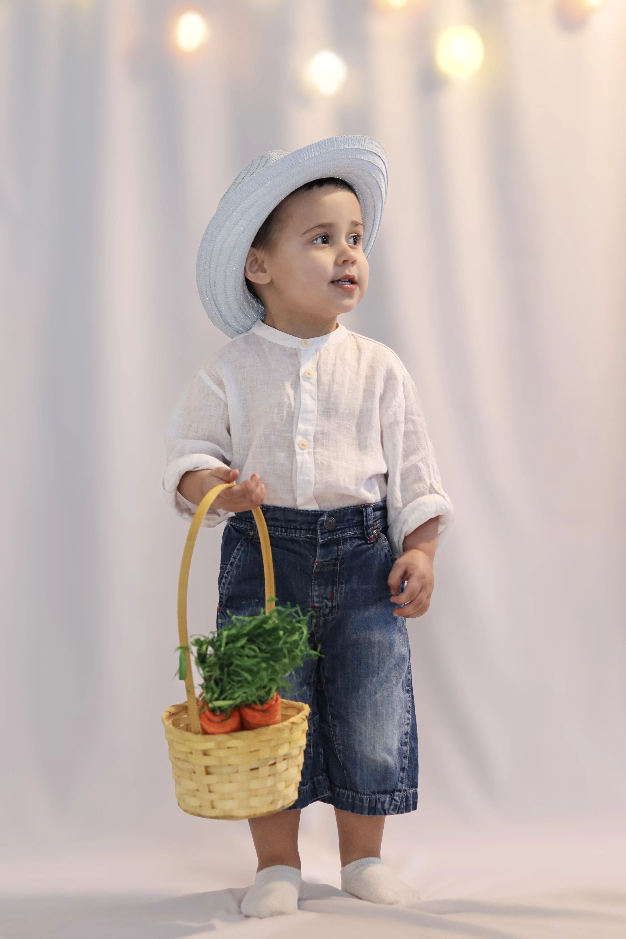 A young child dressed in a white shirt, denim skirt, and white hat holding a basket of carrots and greens, standing against a light-colored background with blurred string lights.