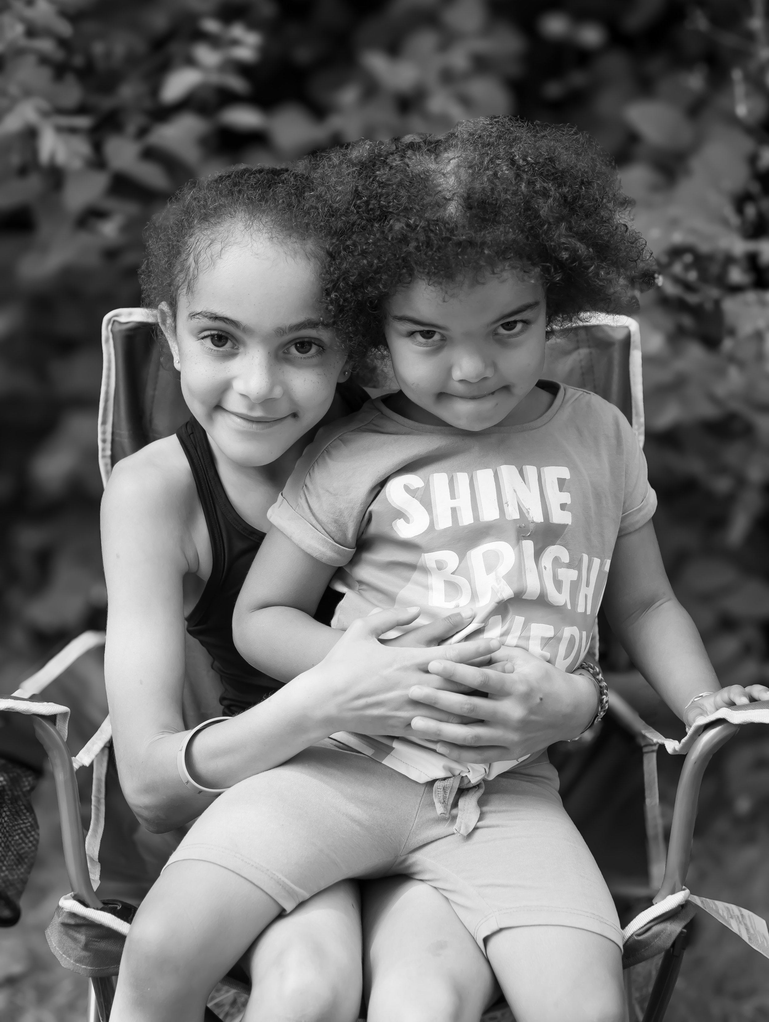 Black and white photo of two young girls sitting together on a chair, smiling and looking at the camera, with a background of blurred foliage.