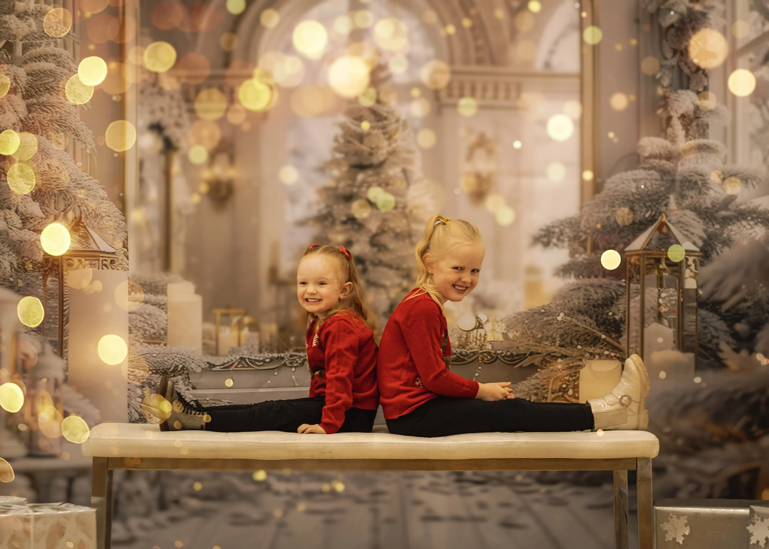 Two young girls in red sweaters and black pants sit back-to-back on a cushioned bench, smiling, with a festive, snowy Christmas scene indoors featuring decorated trees, gift boxes, and warm lights in the background.