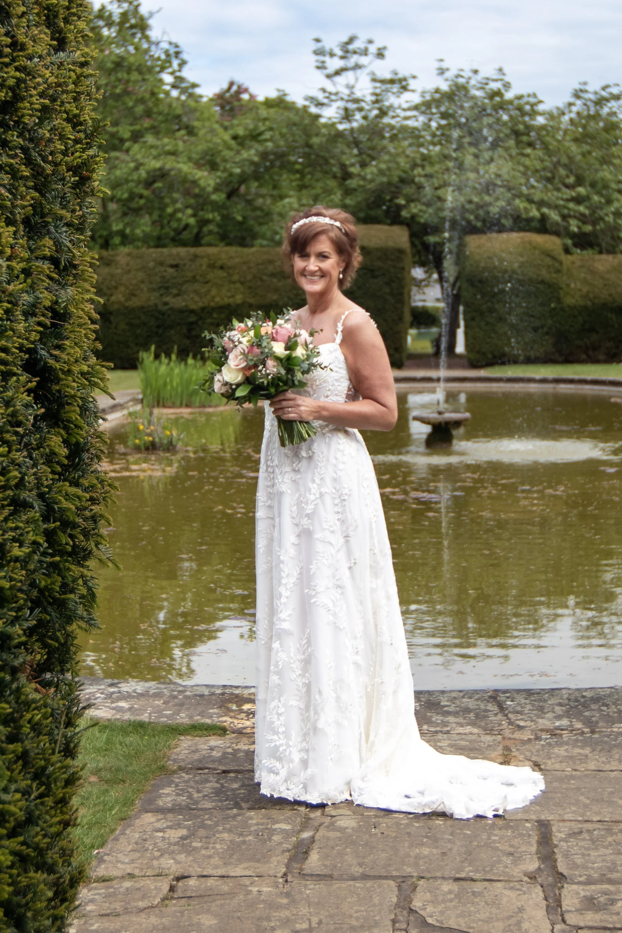 A woman in a white wedding dress holding a bouquet of flowers, standing outdoors near a pond and fountain, with trees and trimmed hedges in the background.