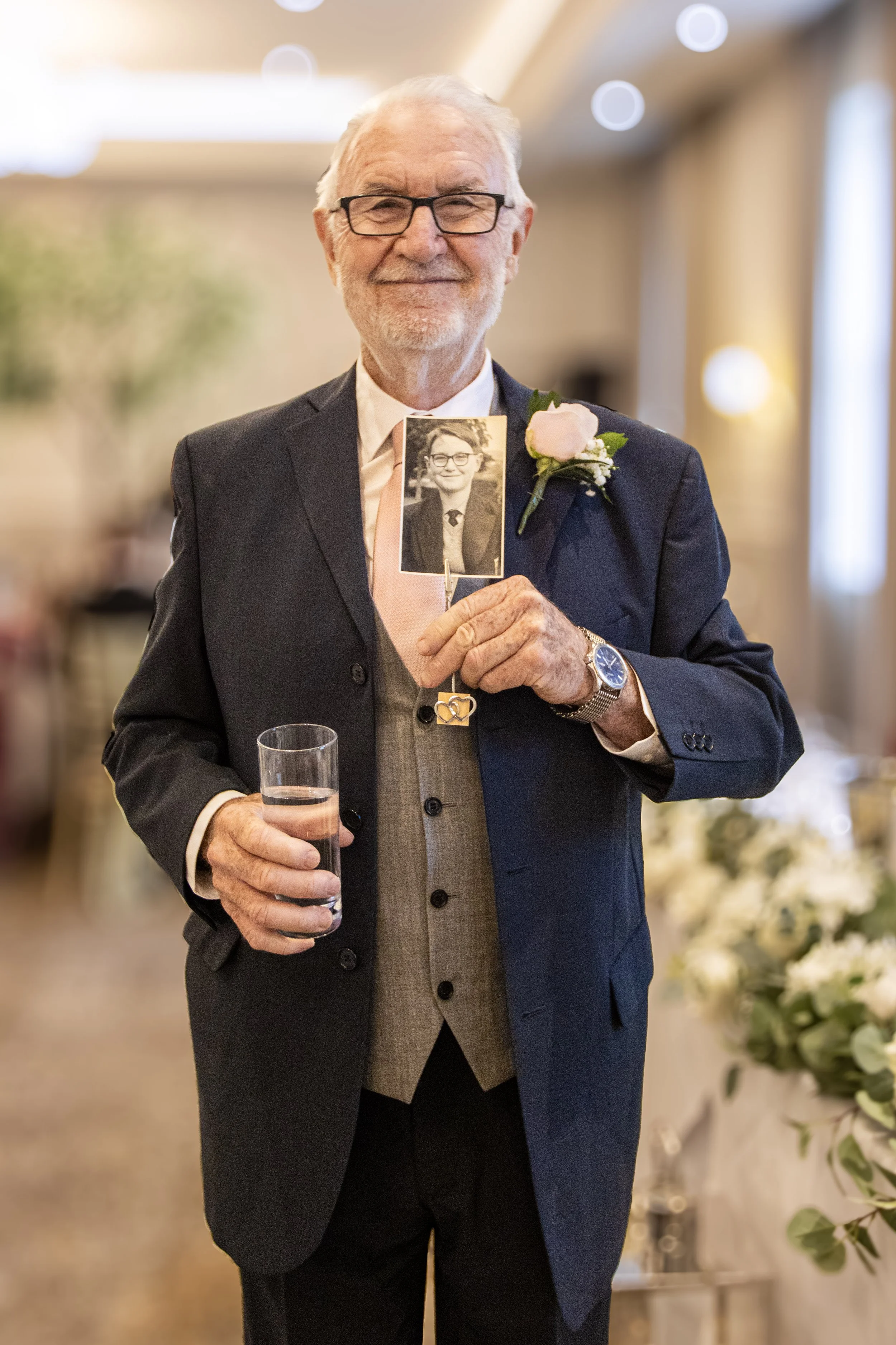 An elderly man in a suit holding a glass of water and a photo of himself as a young man at a formal event.