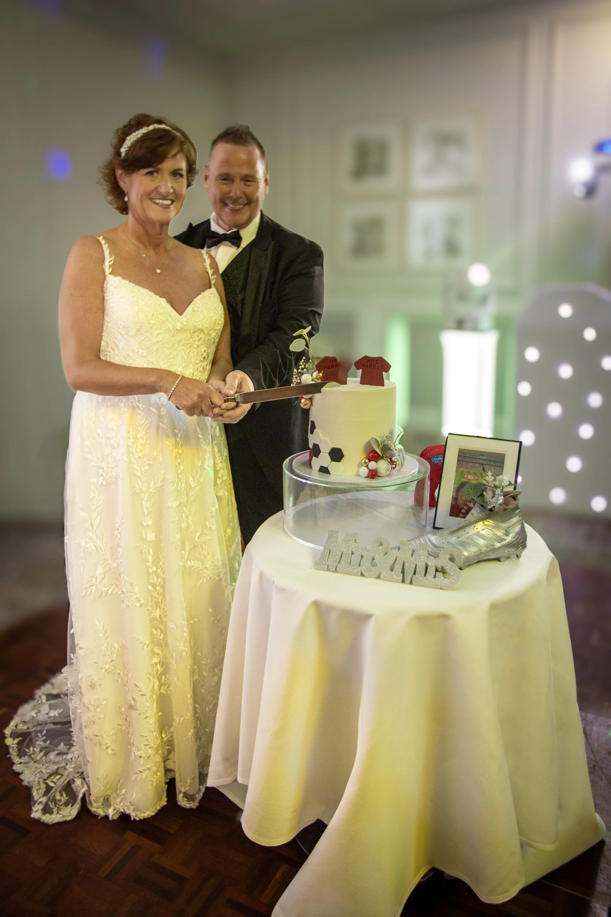 A bride and groom cutting a wedding cake at their reception, standing behind a round table with a white tablecloth. The bride is wearing a yellow gown and a pearl headband, and the groom is in a black tuxedo with a bow tie. The background includes framed photos and decorative lighting.
