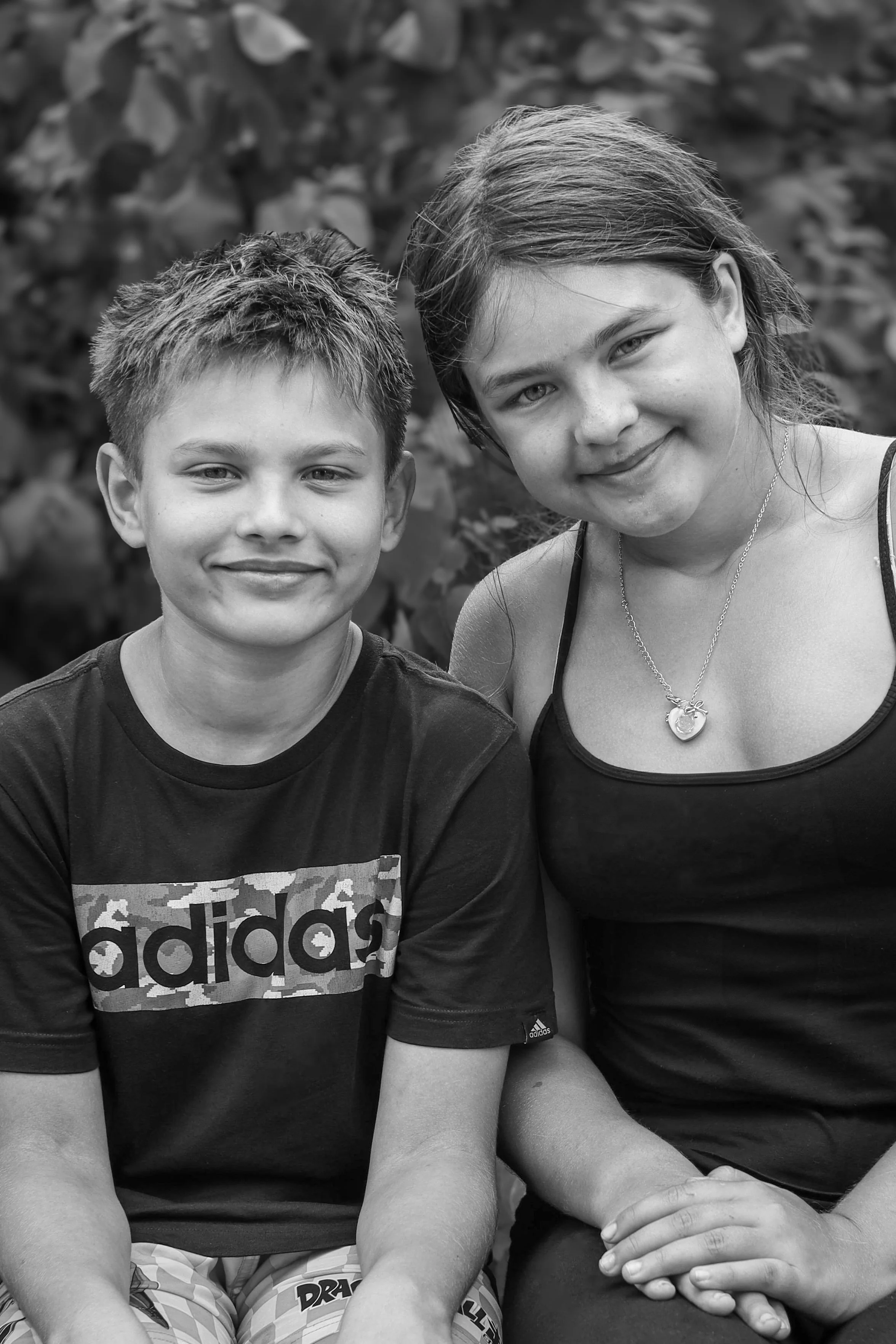 A black and white photo of a smiling teenage boy and girl sitting outdoors, with bushes in the background.