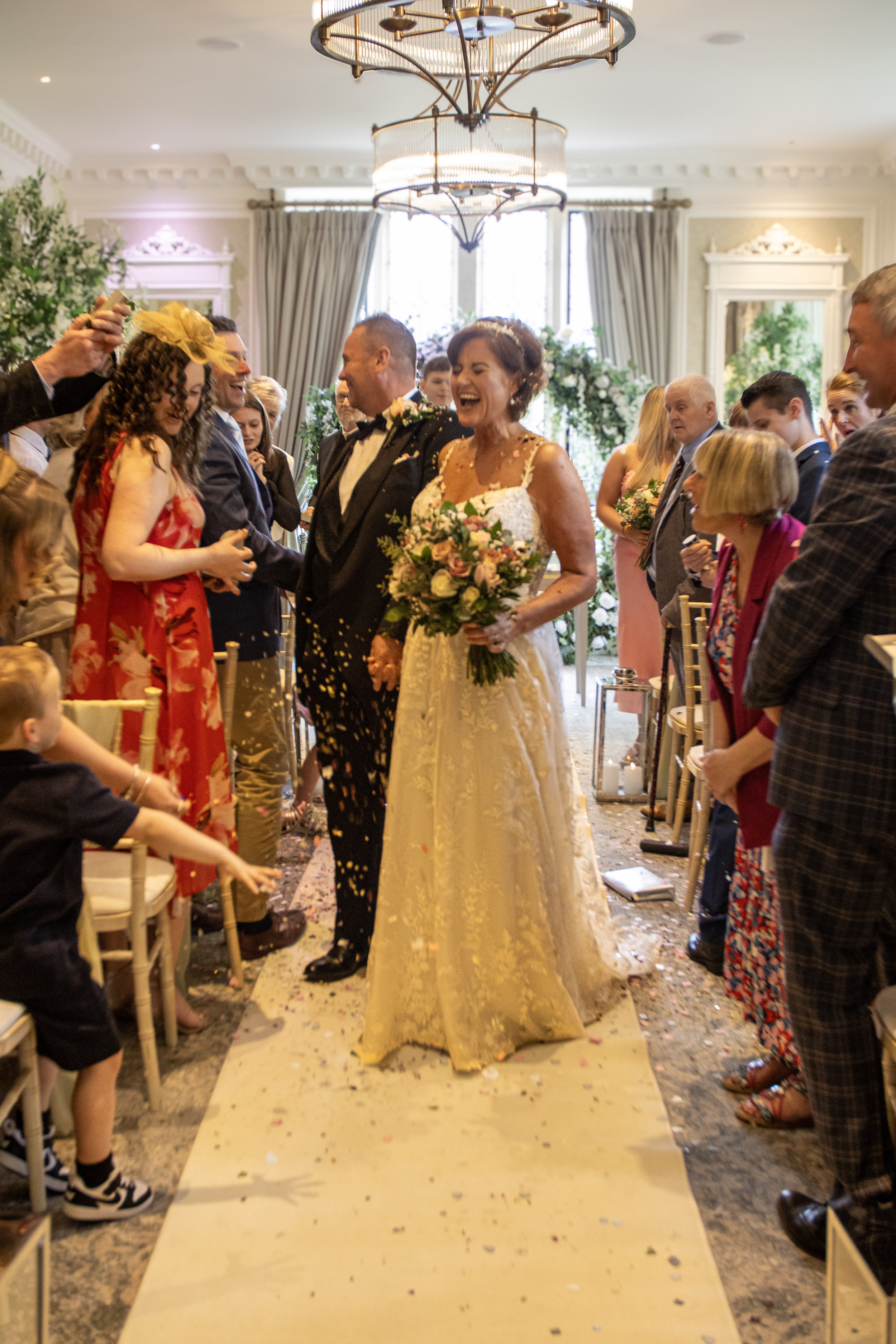 A bride and groom walking down the aisle at their wedding, surrounded by smiling guests, with confetti falling around them in a decorated indoor venue.