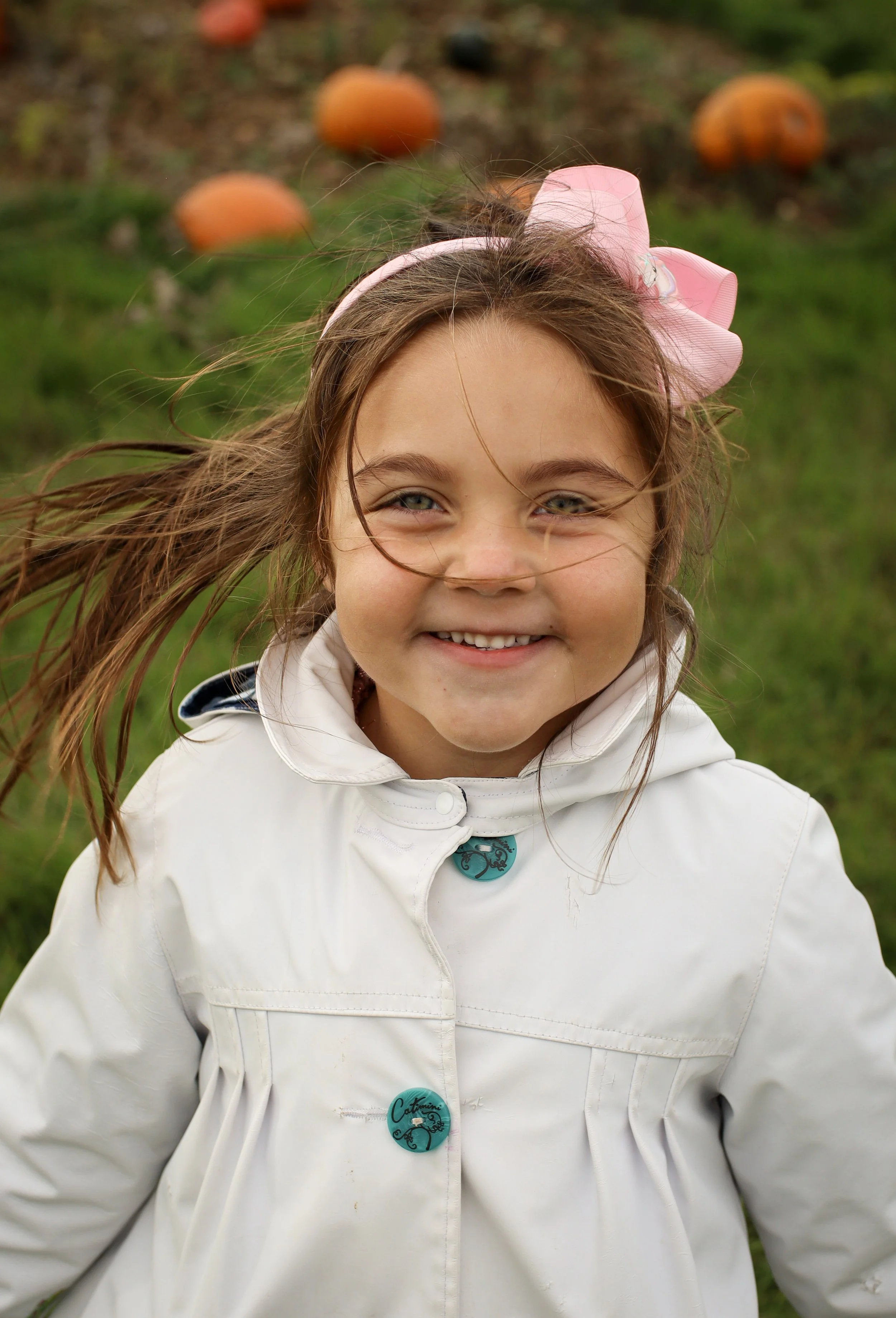 A young girl smiling outdoors, wearing a white jacket with turquoise buttons and a pink bow in her hair, with pumpkins in the background.