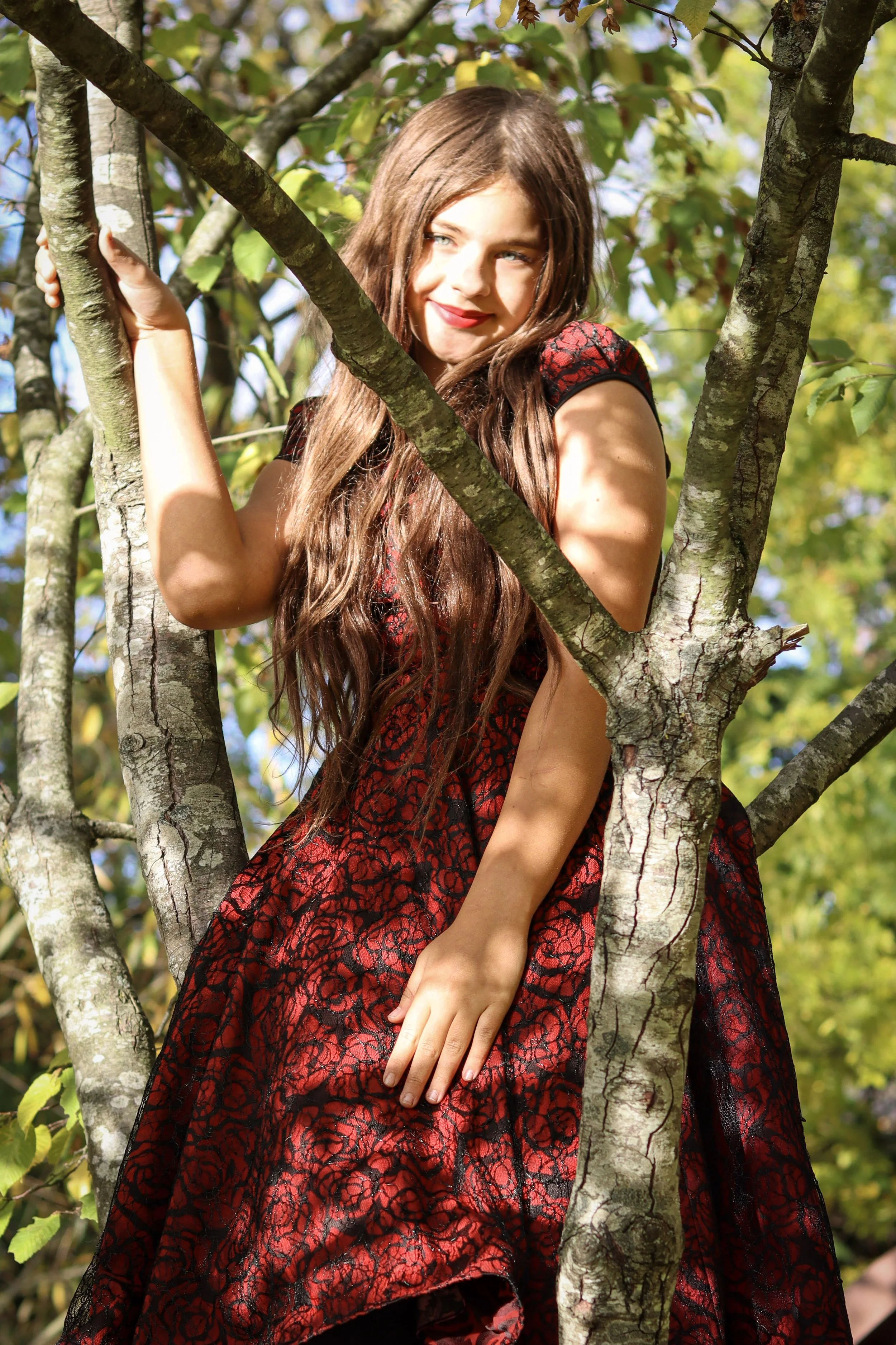 A young woman with long brown hair wearing a red and black patterned dress sitting in a tree, surrounded by green leaves, with sunlight filtering through.