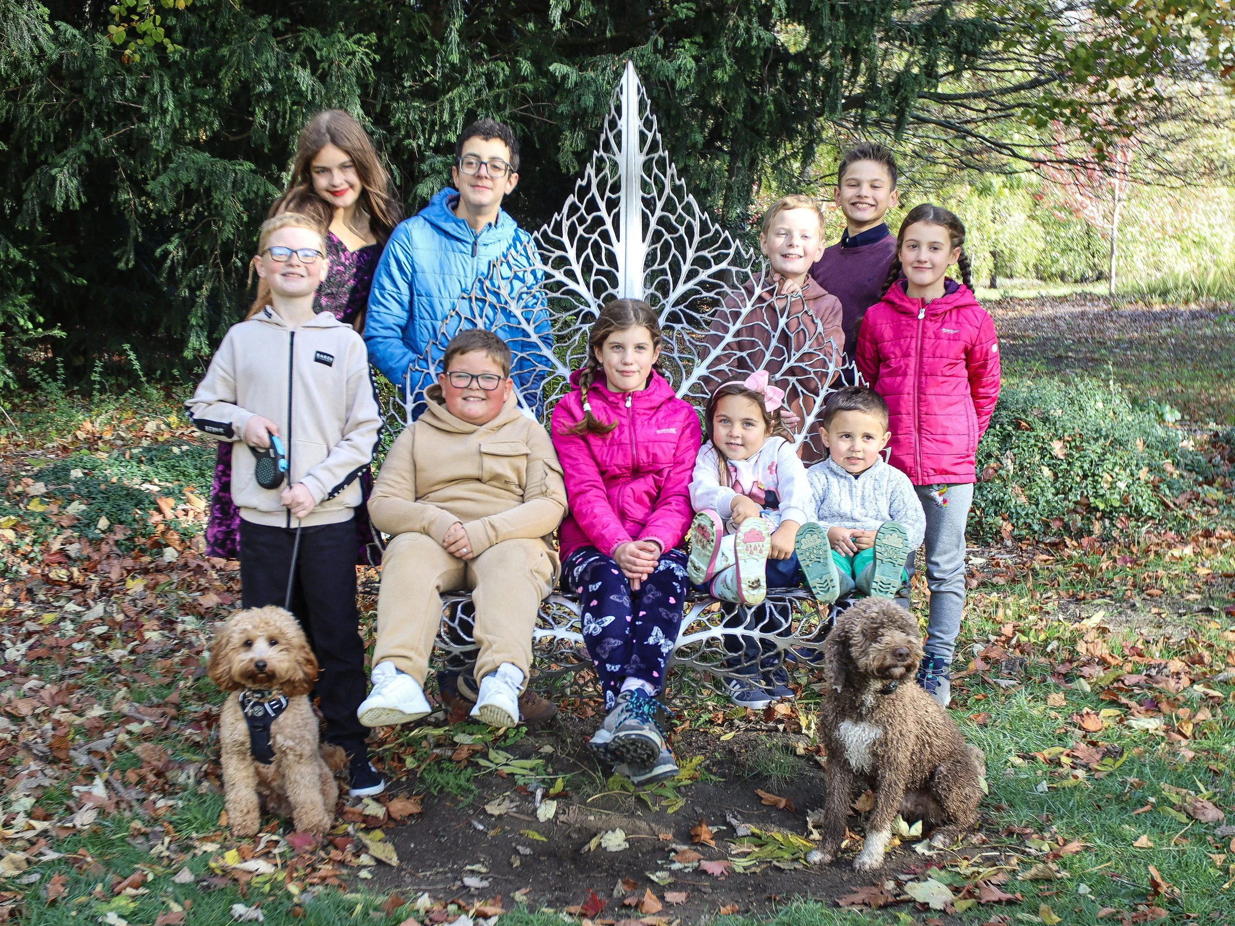 A group of eleven children and two dogs sitting and standing around a decorative bench outdoors in a park during fall. Background shows trees with colorful foliage.