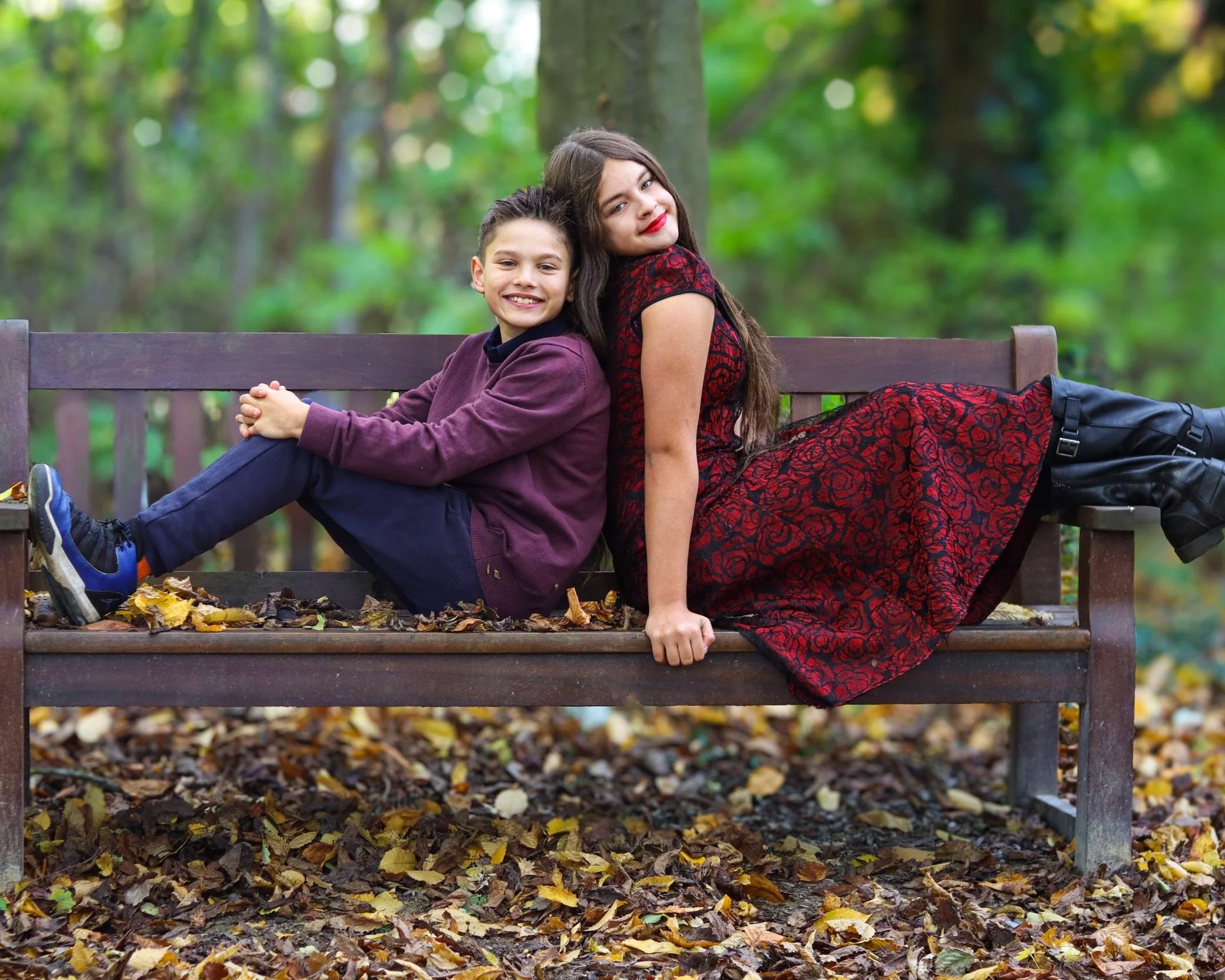 Two children sit on a park bench back-to-back, smiling, surrounded by autumn leaves in a wooded area.