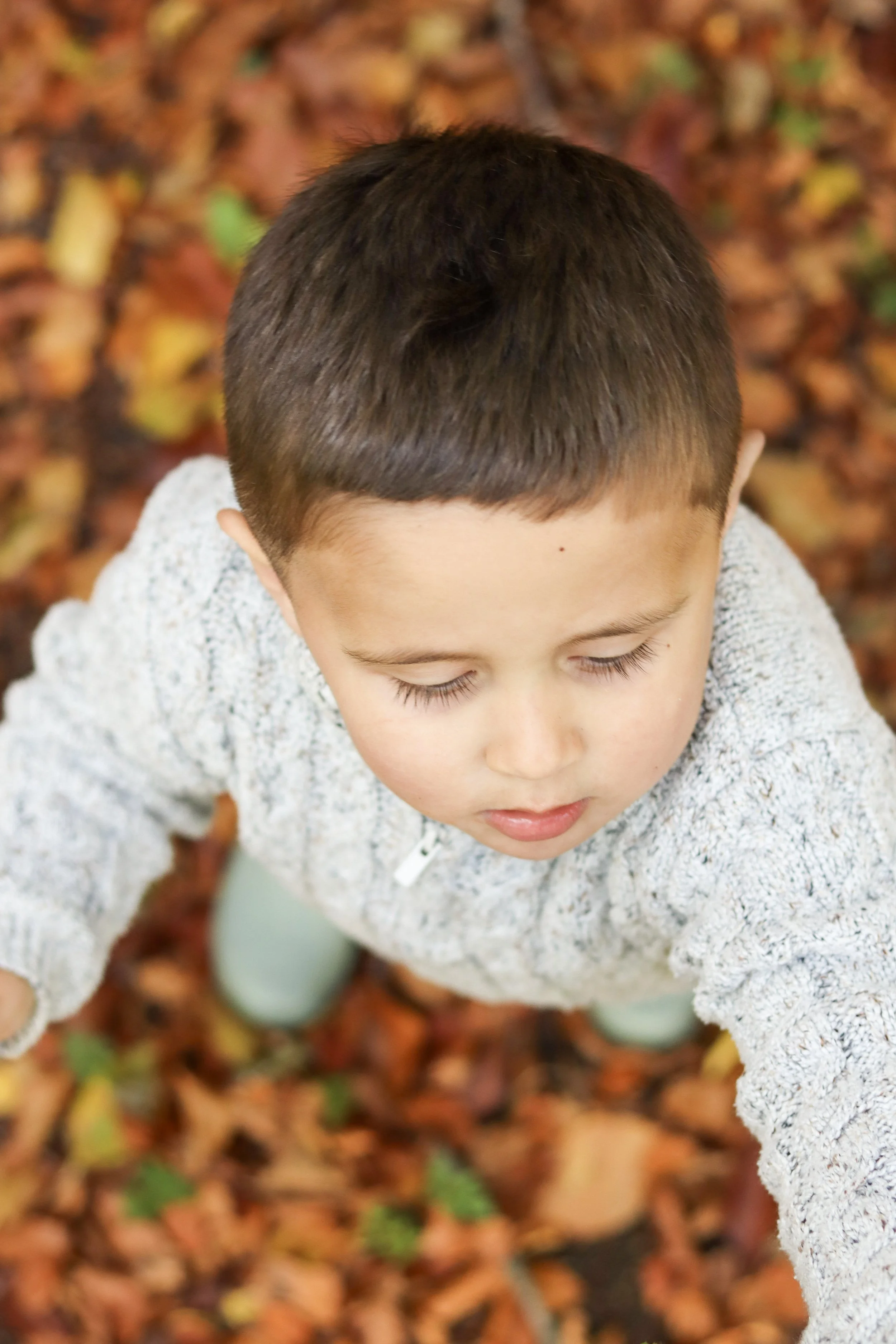 A young boy with short dark hair, closed eyes, and a neutral expression, standing outdoors among fallen autumn leaves.