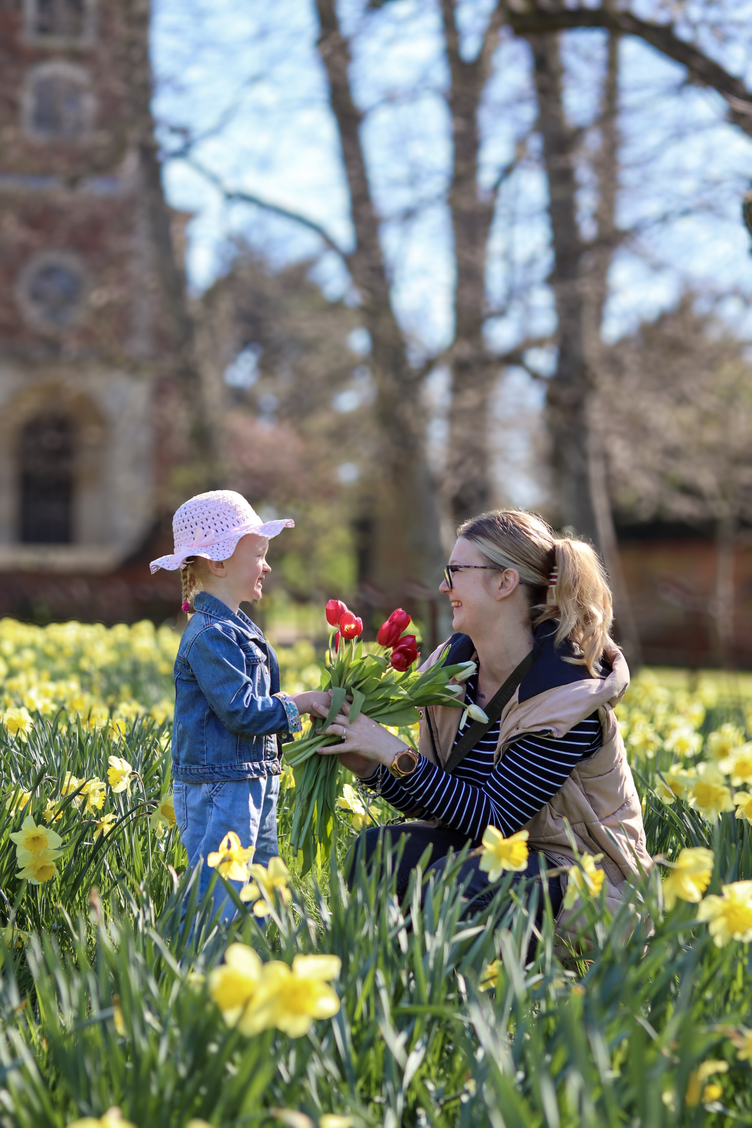 Woman giving a bouquet of red and white flowers to young girl in a flower field with yellow flowers, trees, and a building in the background.
