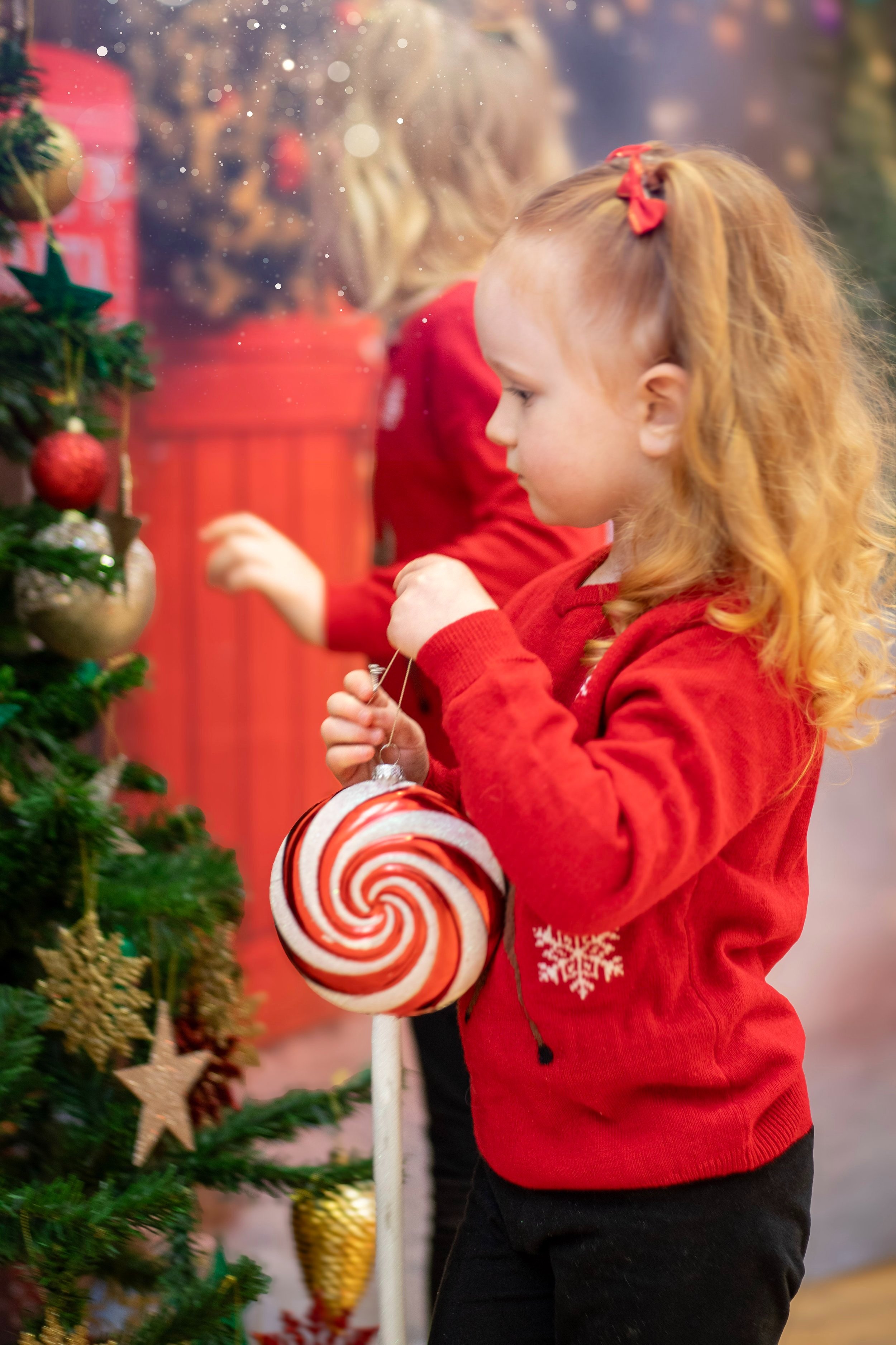 A young girl with red hair, wearing a red sweater, decorating a Christmas tree with a large red and white swirled lollipop ornament.