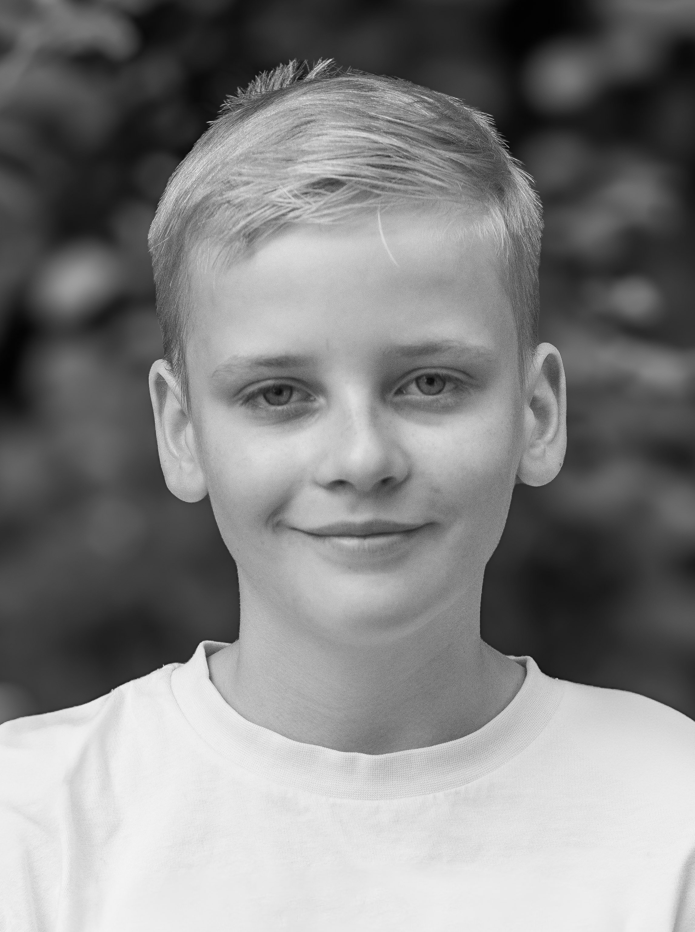 Black and white portrait of a young boy with short light hair, smiling, wearing a light-colored t-shirt, with blurred foliage background.