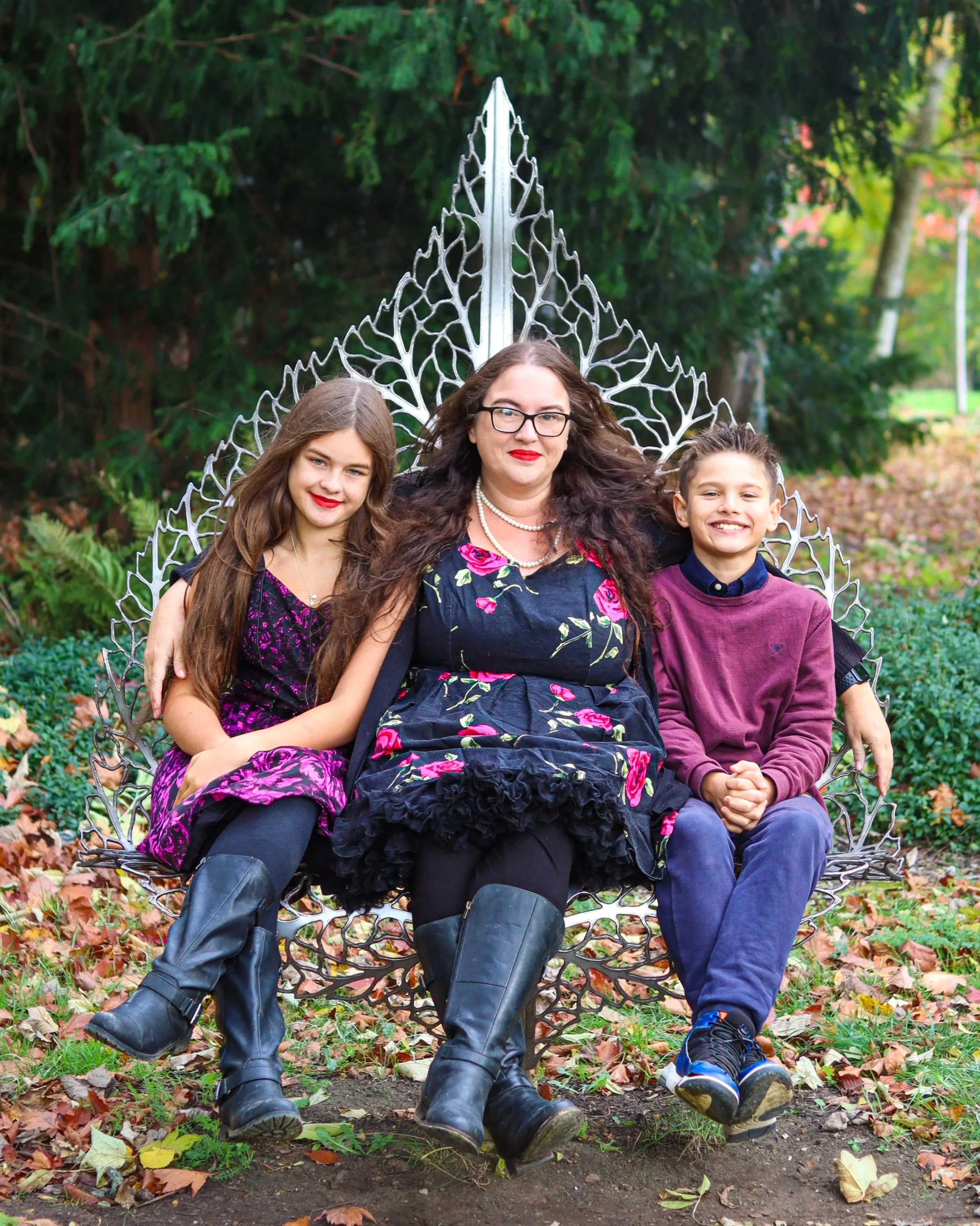 A woman with two children sitting on decorative metal garden bench outdoors, surrounded by greenery and fallen autumn leaves.