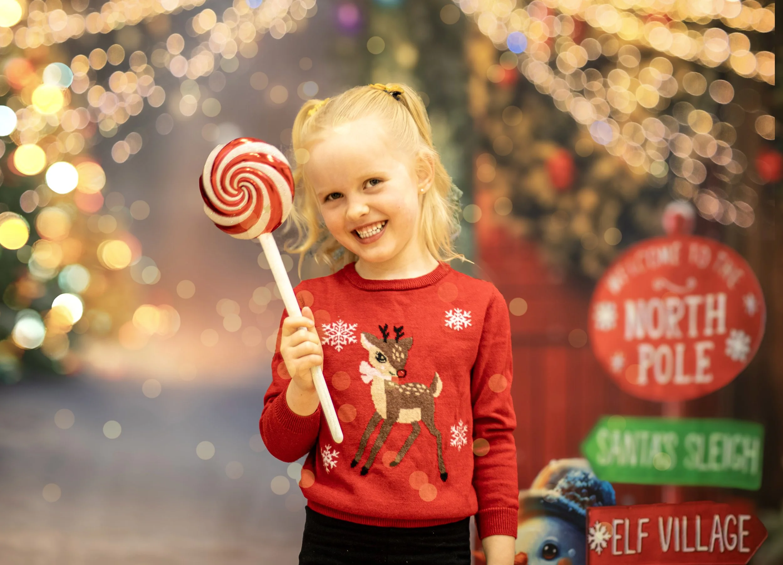 A young girl with blonde hair in pigtails smiling and holding a large red and white swirled lollipop, standing in front of a festive, blurred Christmas background with colorful lights and signs including one that says "North Pole" and another that says "Santa's Sleigh." She is wearing a red Christmas sweater with a reindeer design.