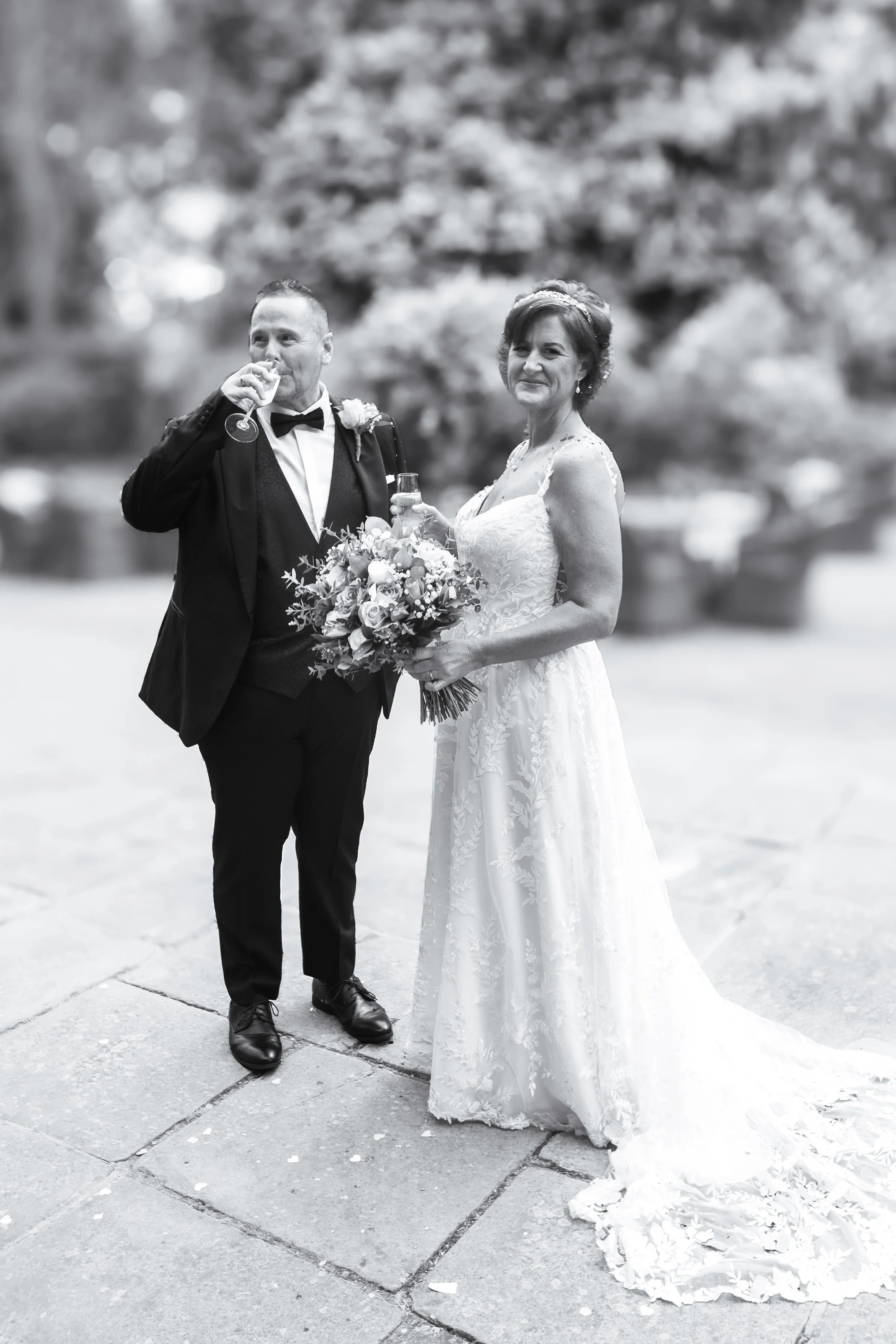 Black and white photo of a bride and groom at their wedding, the groom drinking from a glass and the bride holding a bouquet, standing outdoors on a paved area with trees in the background.