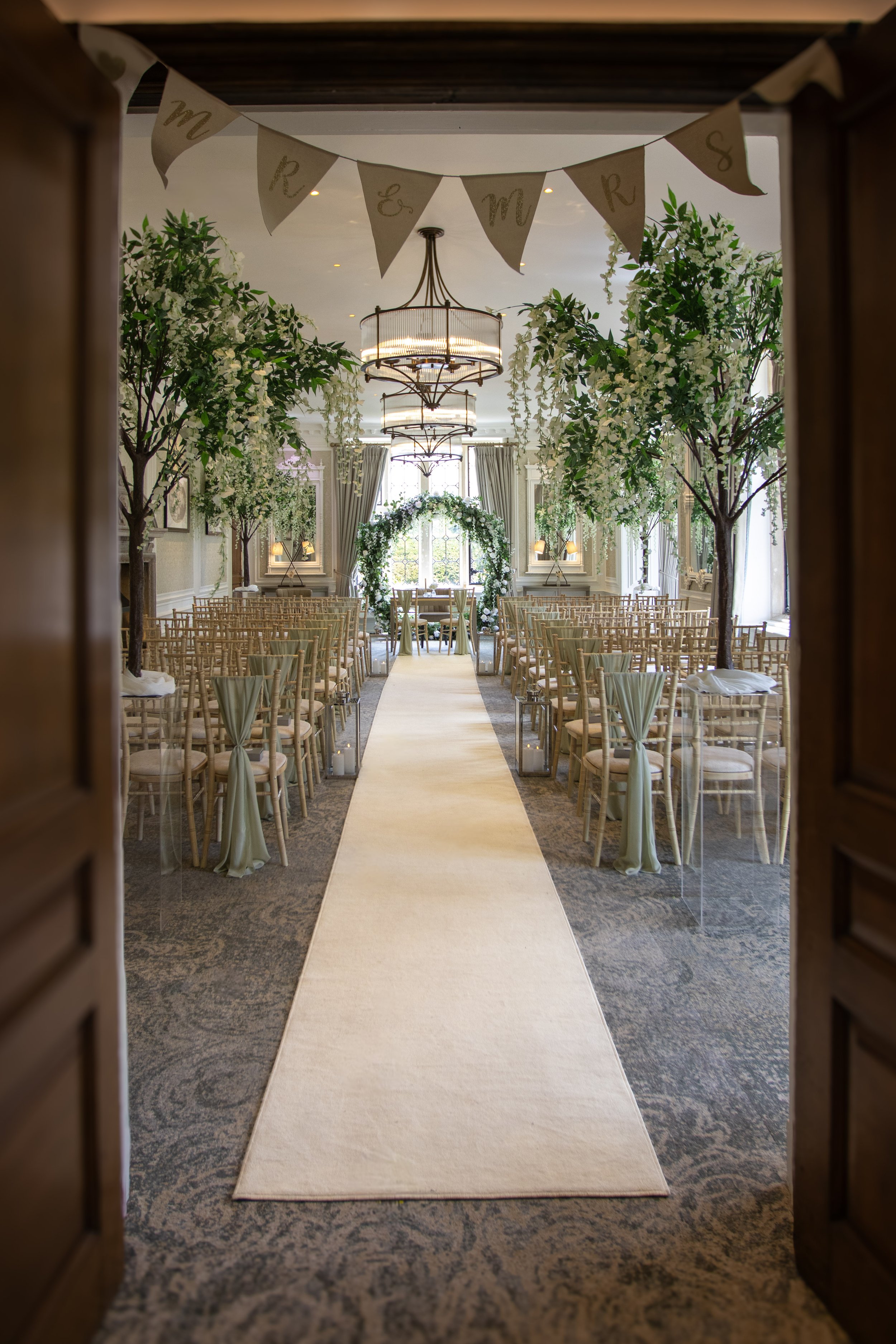 Elegant wedding ceremony setup with chairs, floral trees, an arch decorated with flowers, and a cream-colored aisle runner, viewed through a doorway.