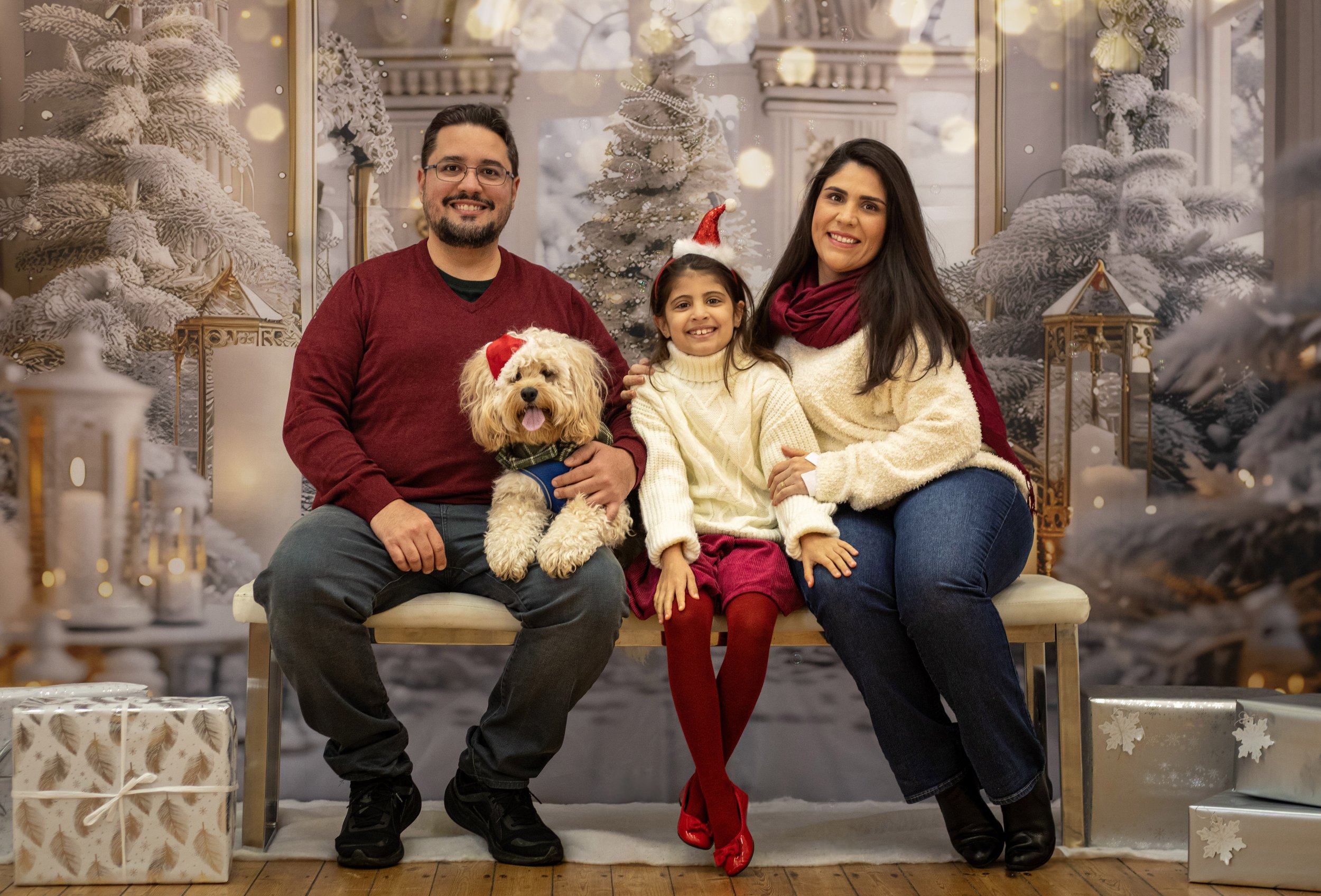 A family of four with a dog sitting on a bench in front of a Christmas-themed background, including decorated trees and holiday decorations, with presents at their feet, celebrating the holidays.