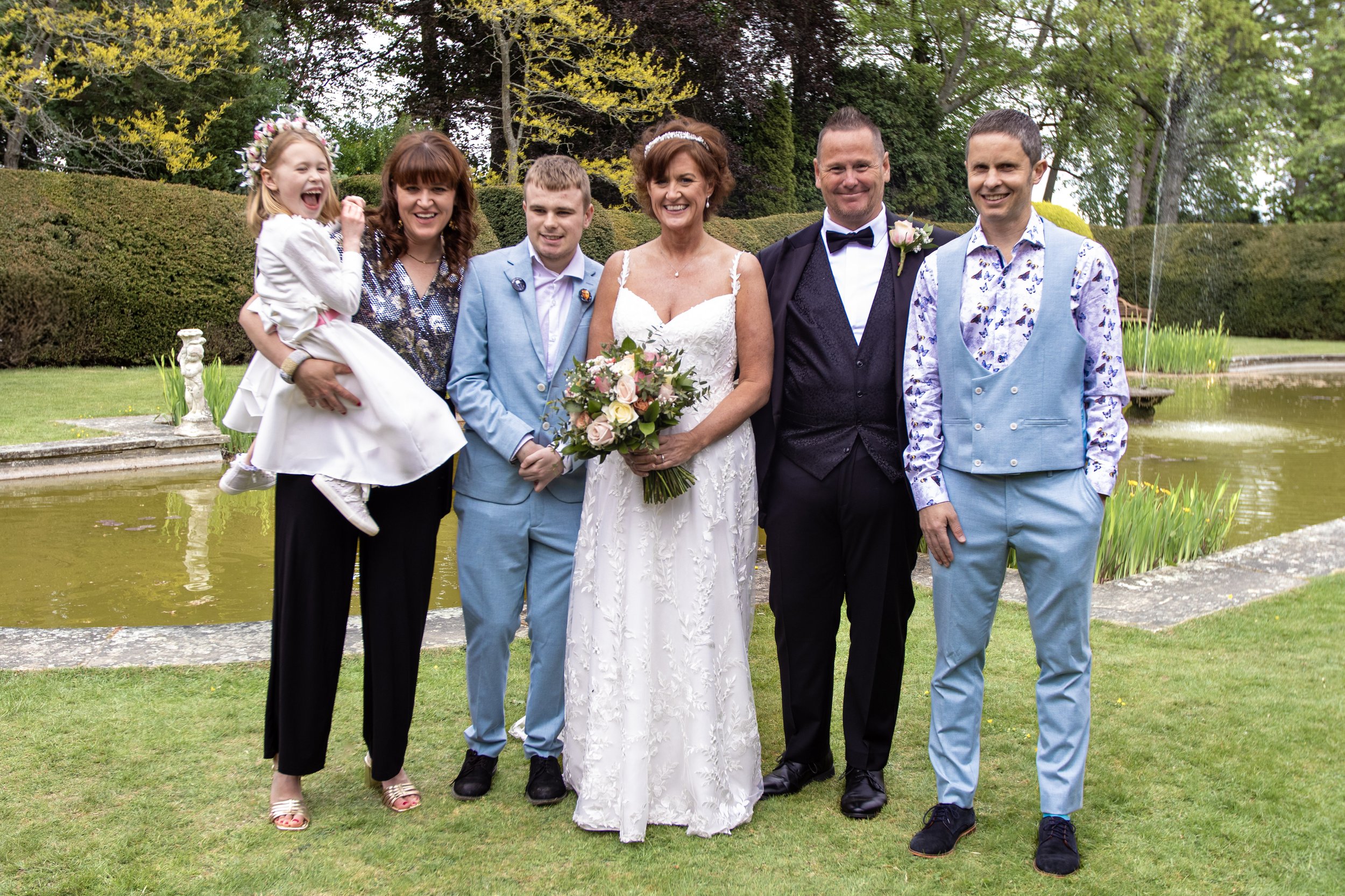A group of seven people, including a bride in a white wedding dress holding a bouquet, and groom in a black tuxedo, standing outdoors near a pond, surrounded by greenery and trees.