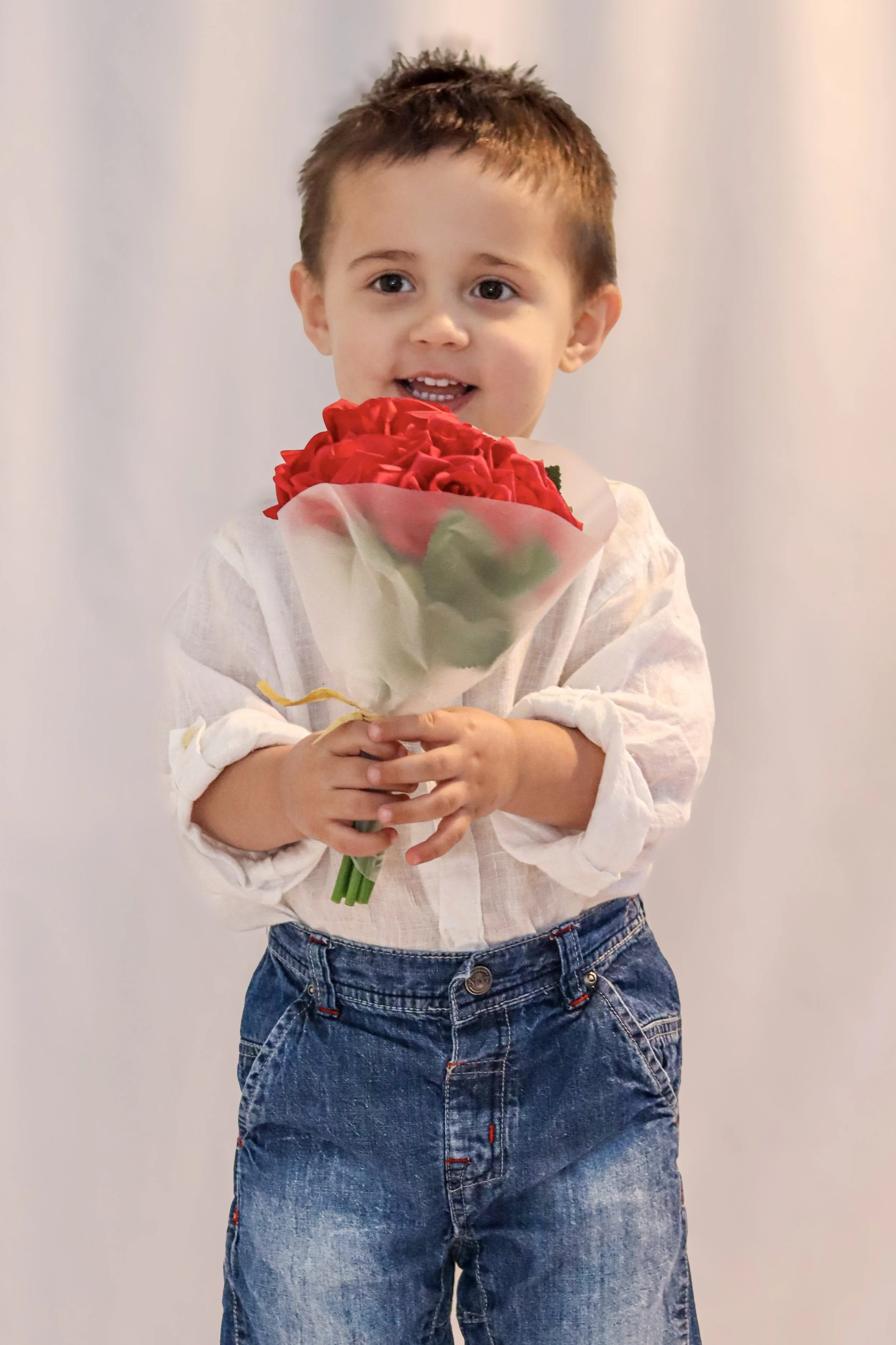 A young boy with short brown hair wearing a white shirt and blue jeans holding a bouquet of red roses and smiling.