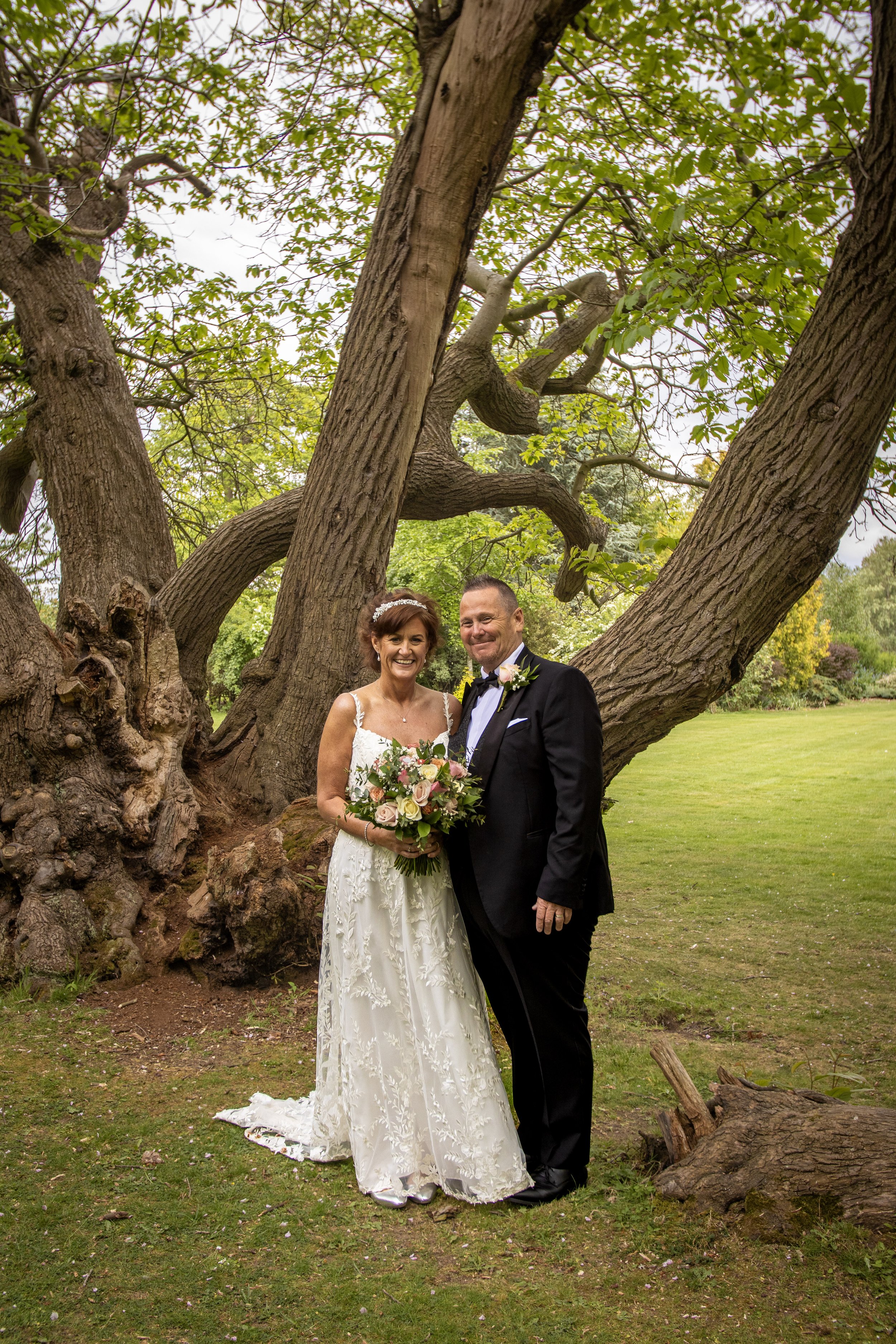 Bride and groom standing together outdoors in front of a large, twisted tree, smiling, dressed in wedding attire. The bride wears a white lace wedding gown and holds a bouquet of flowers, while the groom wears a black tuxedo with a white shirt and bow tie.