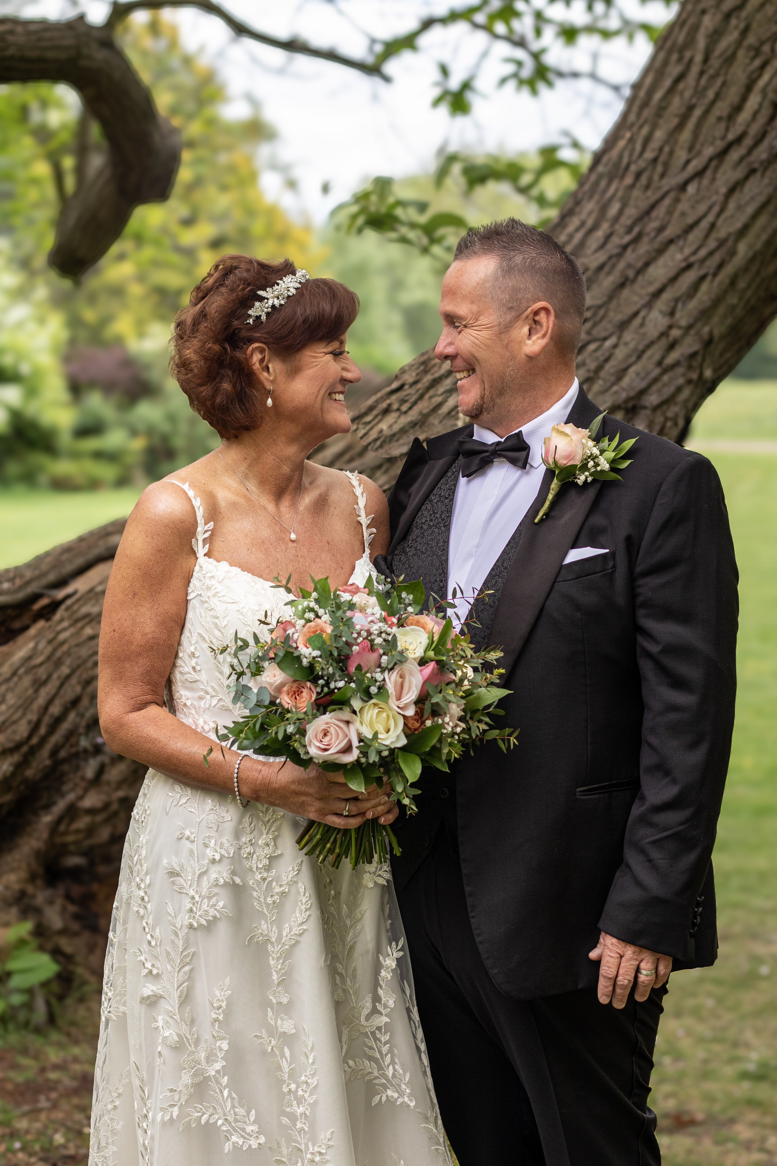 A smiling bride and groom outdoors near a large tree, holding a floral bouquet, dressed in wedding attire.