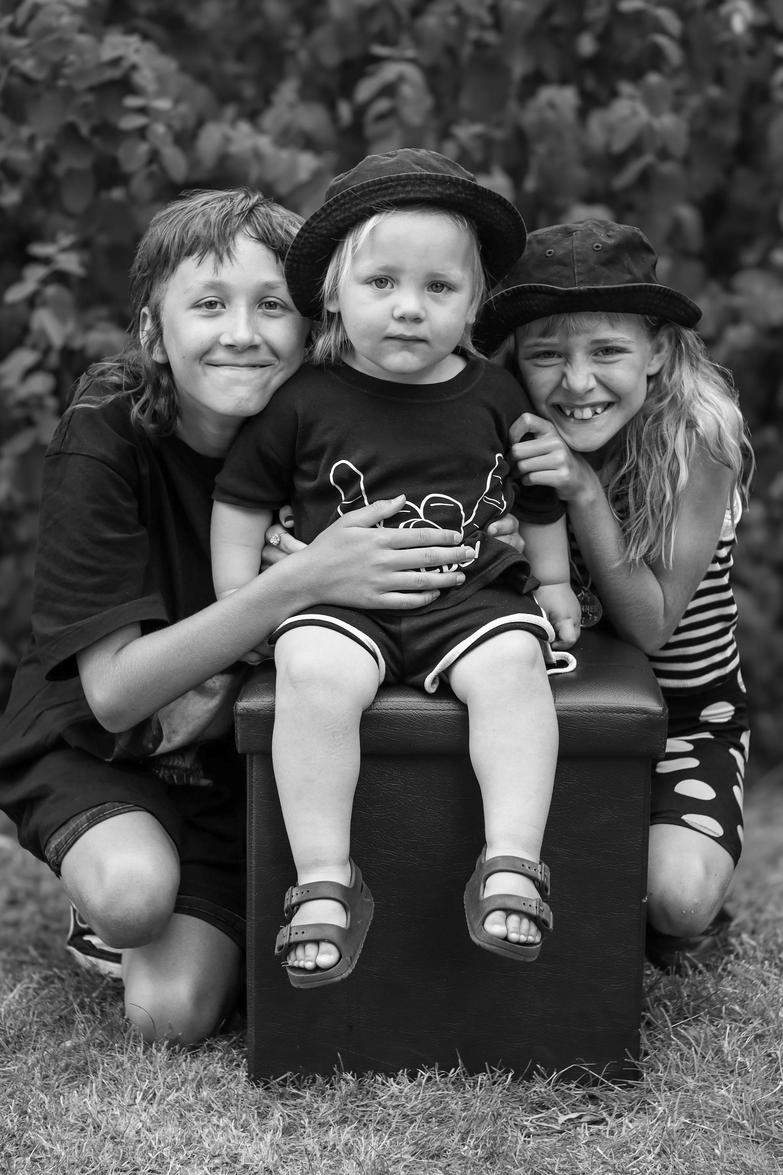 Three children outdoors, sitting on a box, smiling and posing for the photo. Two older children on each side have hat accessories and are hugging the youngest child in the middle, who is sitting and looking serious.