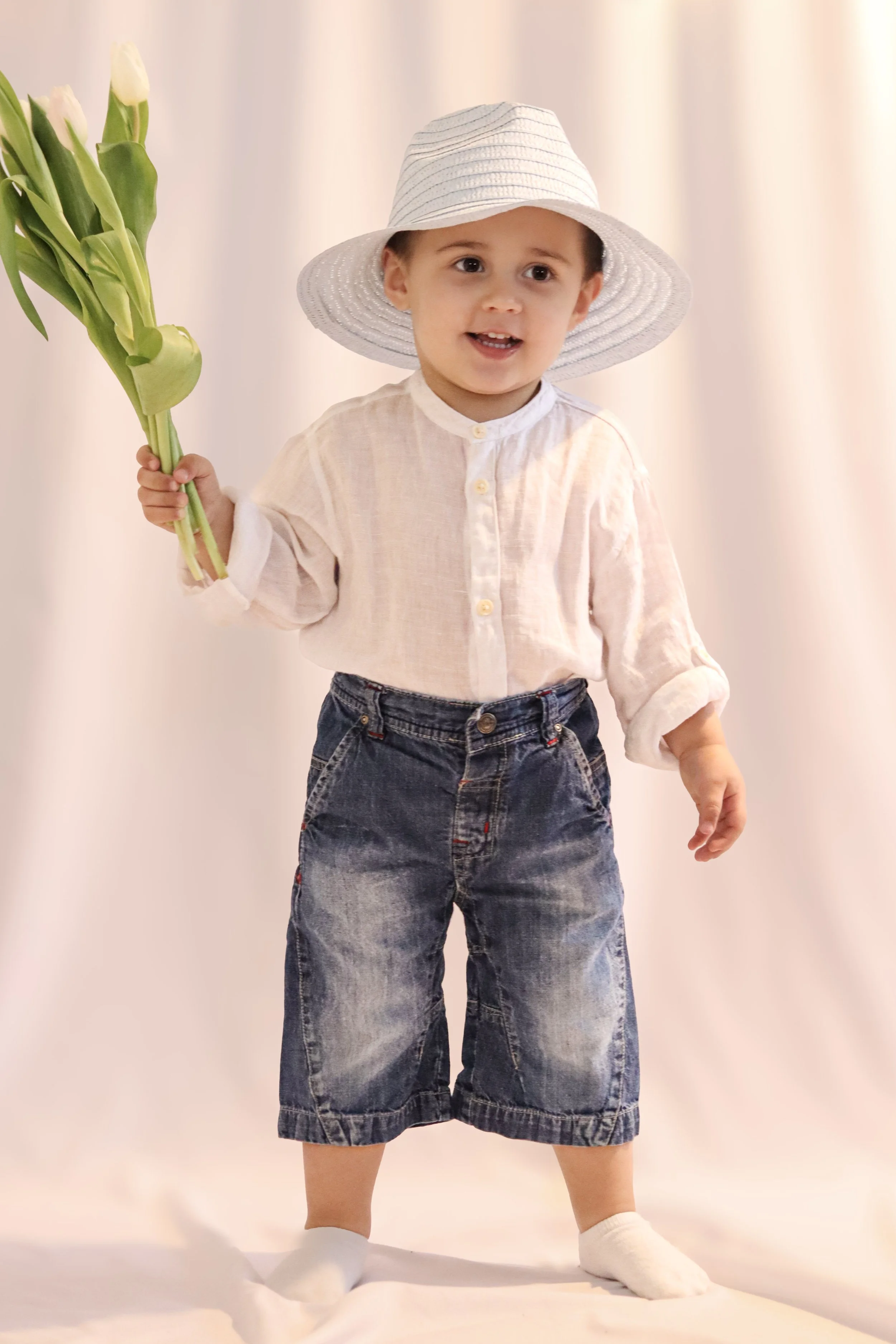 A young boy wearing a white shirt, denim shorts, a sun hat, and white socks holding a bouquet of white tulips.