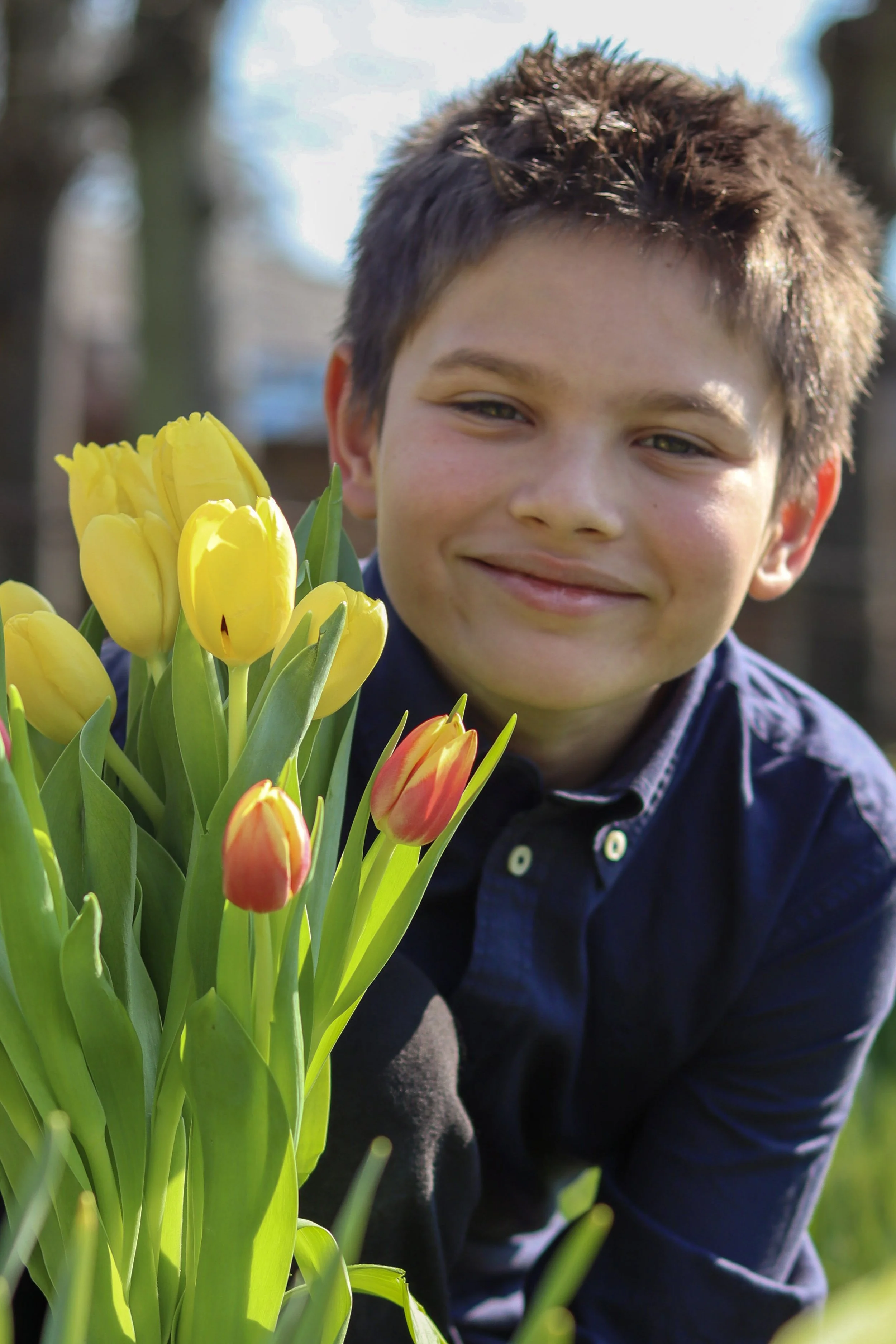 A smiling young boy with short brown hair holding a bouquet of yellow and orange tulips outdoors on a sunny day.