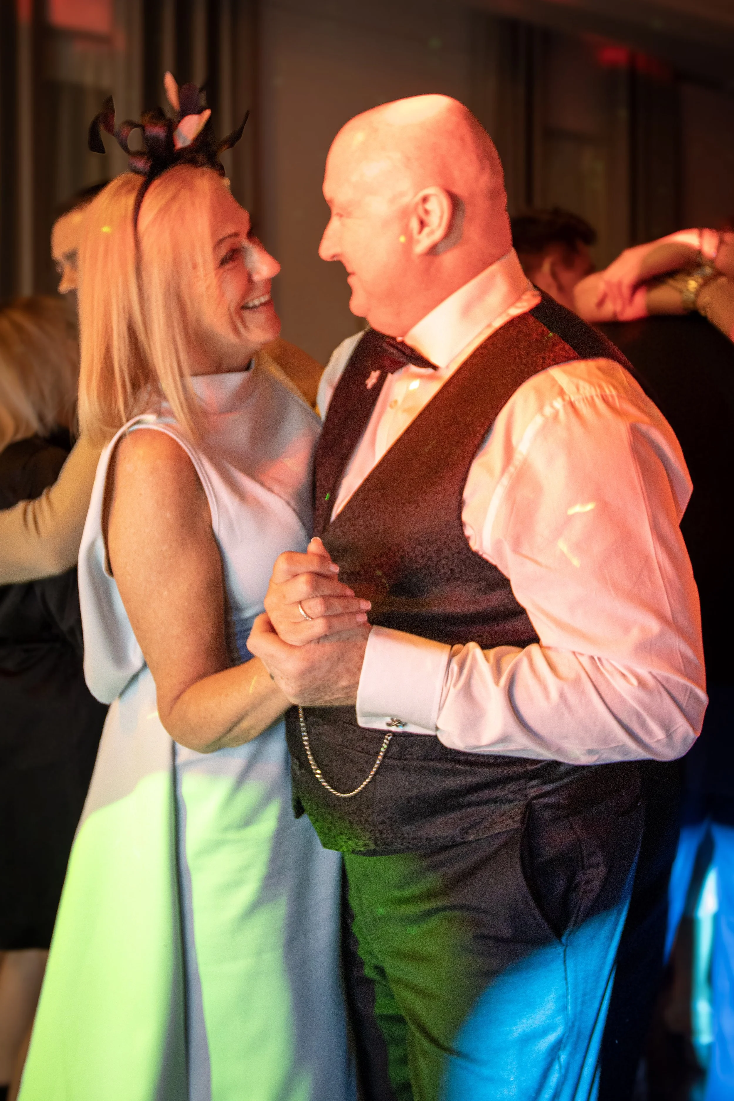 A couple dancing closely and smiling at each other at an indoor event, with colorful lighting and other people in the background.