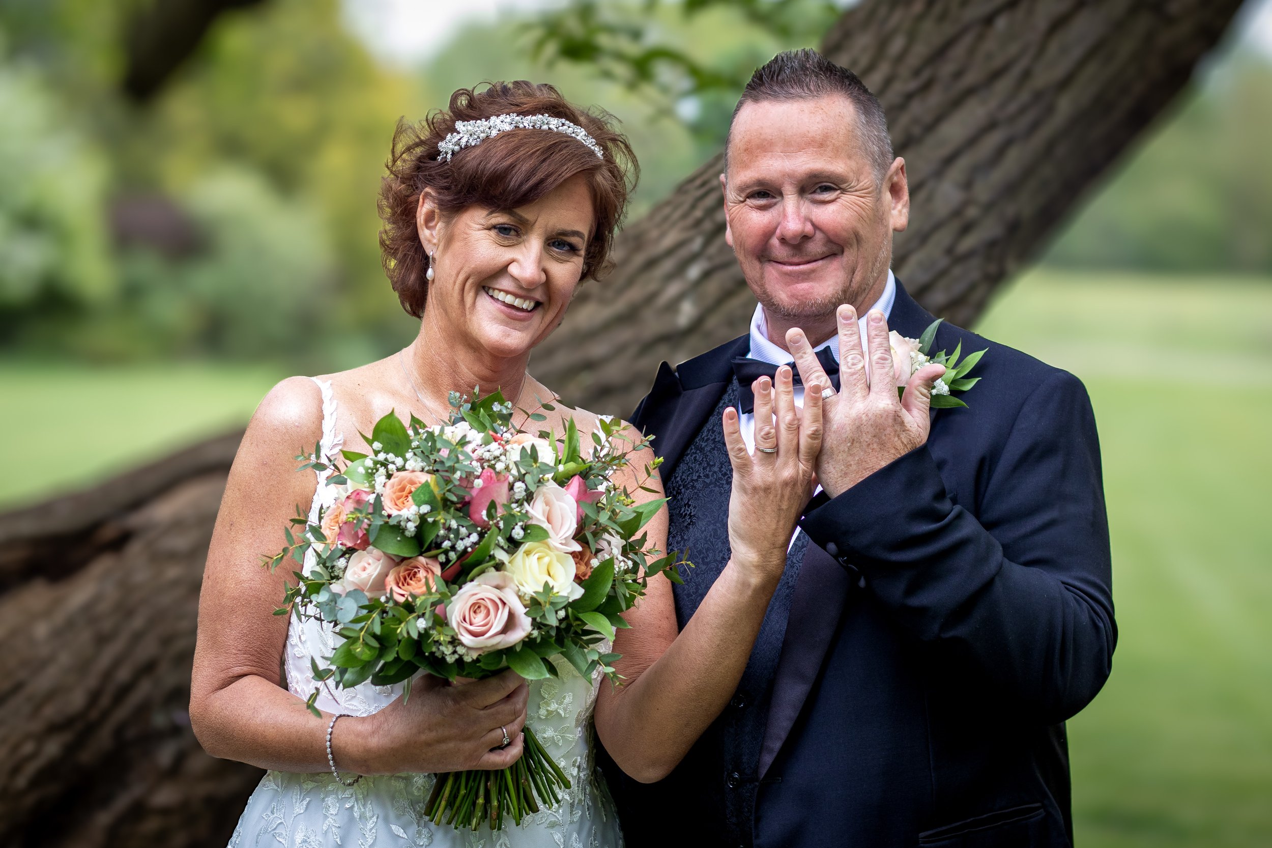 A smiling bride in a white wedding dress and a groom in a black tuxedo standing outdoors in front of a large tree, holding a bouquet of pink, white, and peach roses, with the groom showing his wedding band.