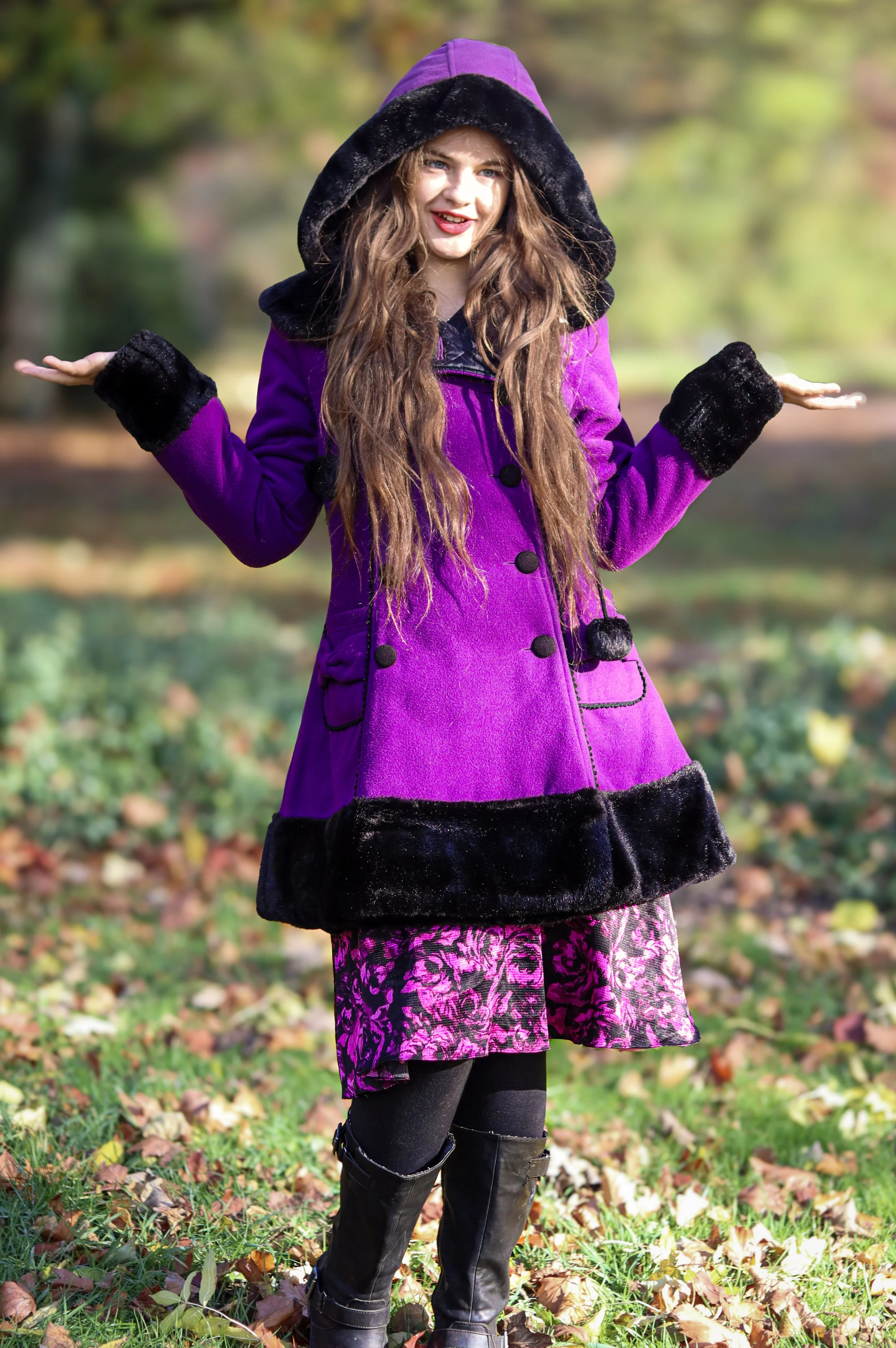 A woman standing outdoors on fallen autumn leaves, wearing a purple coat with black fur trim, a patterned skirt, and tall black boots, with her arms raised and hands open.