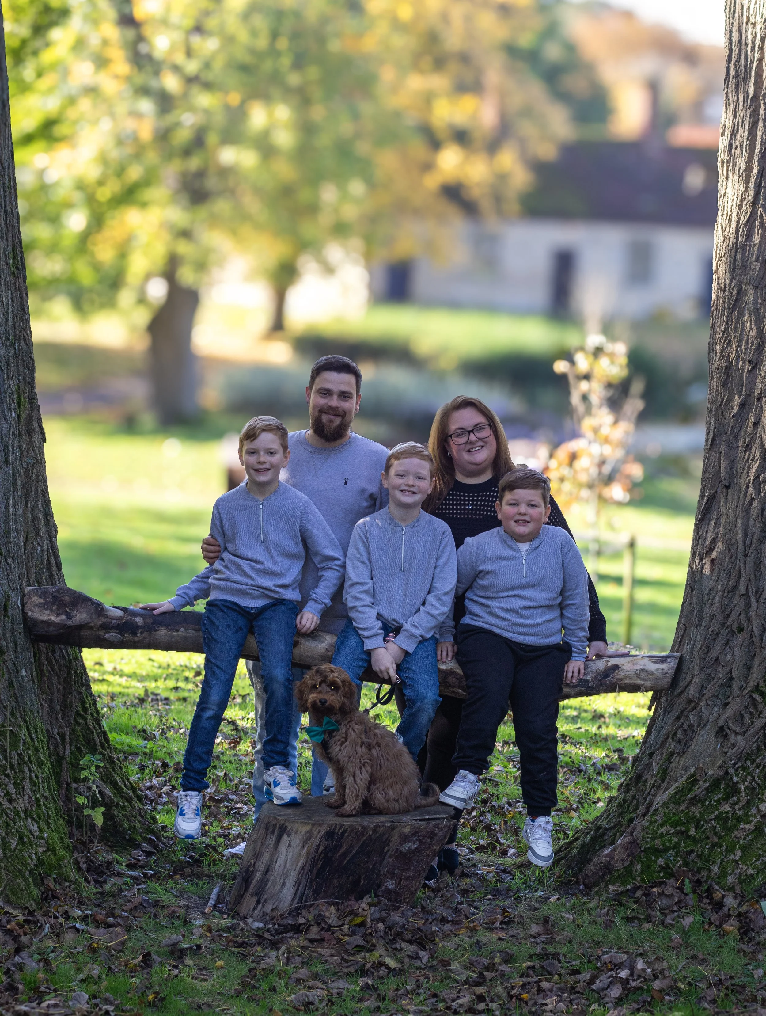 Family of five with a dog posing outdoors between two trees, sitting on a branch, with a house and trees in the background.