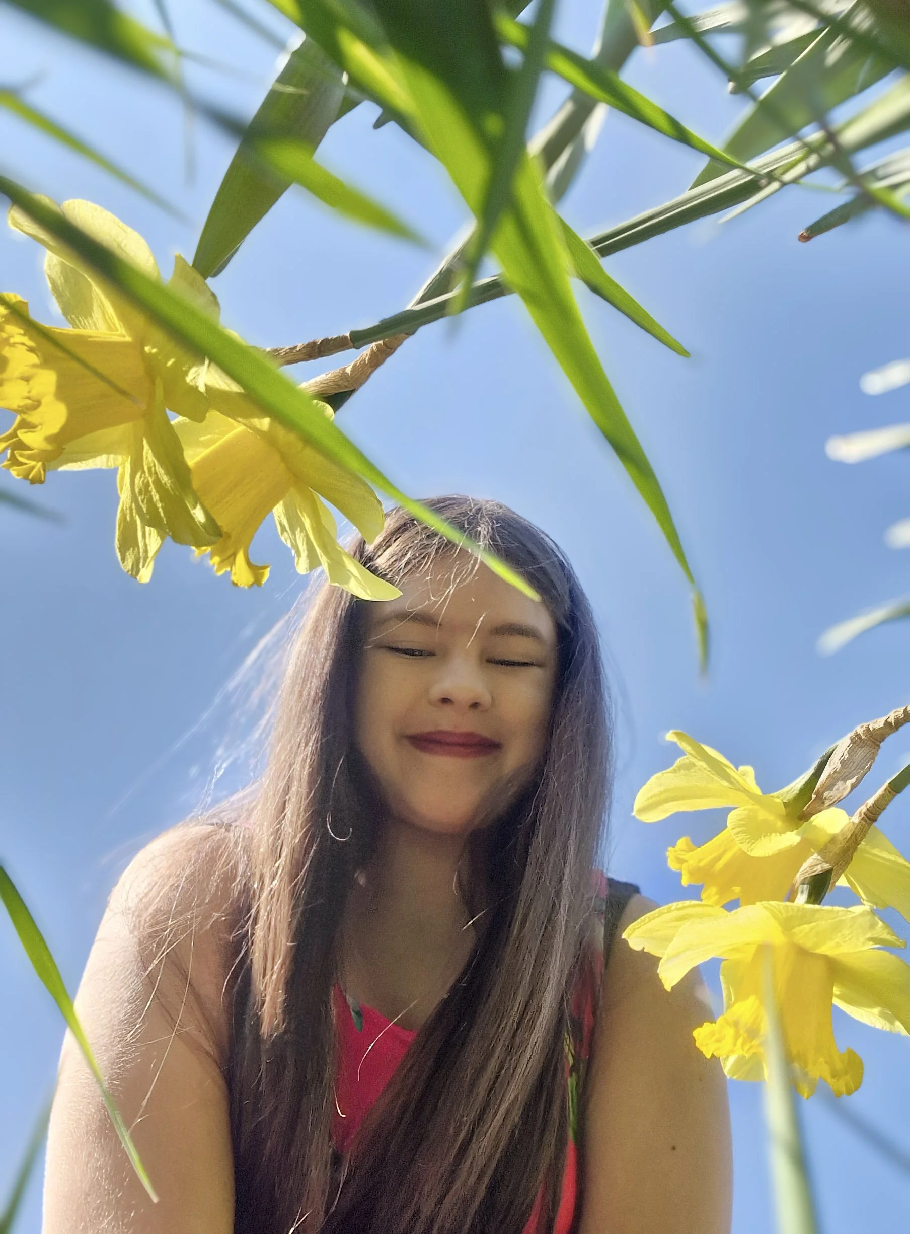 A young woman with long dark hair and a red top smiling with eyes closed, surrounded by yellow flowers and green leaves, under a bright blue sky.