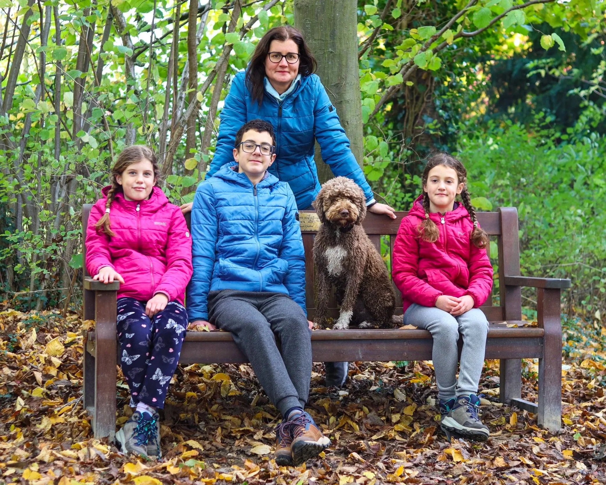 A woman and three children sitting on a park bench with a dog, surrounded by trees and fallen autumn leaves.
