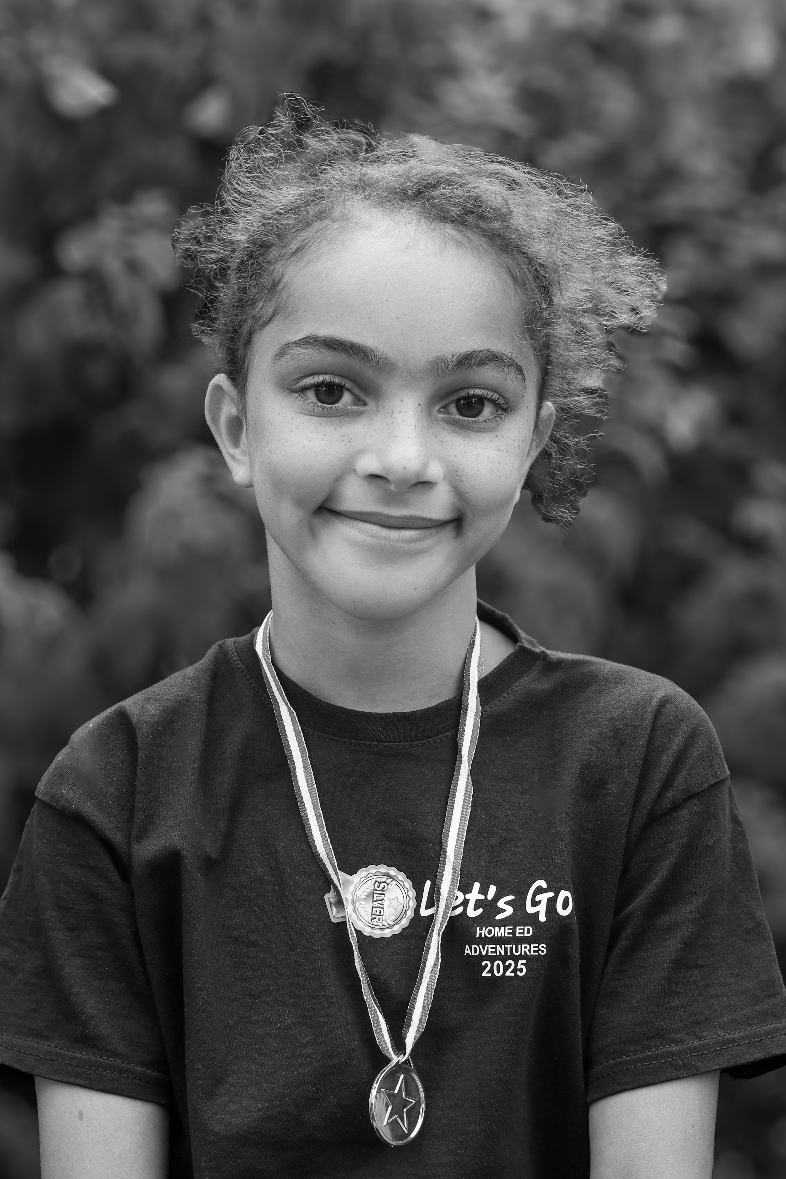 A young girl with curly hair and freckles, smiling, wearing a t-shirt with the text 'Let's Go Home Ed Adventures 2025,' and sporting two medals around her neck, one silver and one bronze, with a blurred natural background.