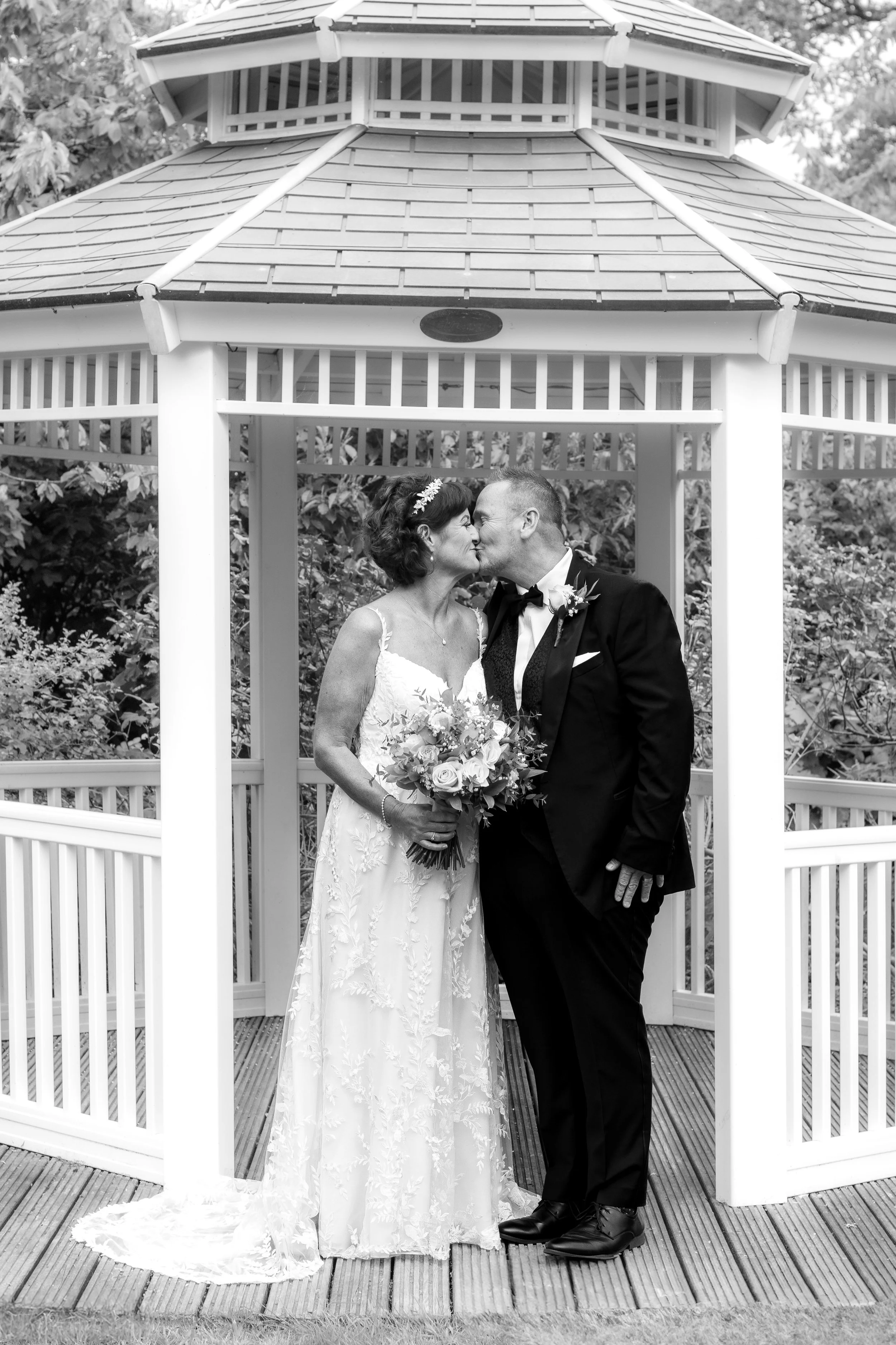 Black and white photo of a bride and groom sharing a kiss in a gazebo. The bride is holding a bouquet and wearing a lace wedding dress, while the groom is in a tuxedo with a bowtie.