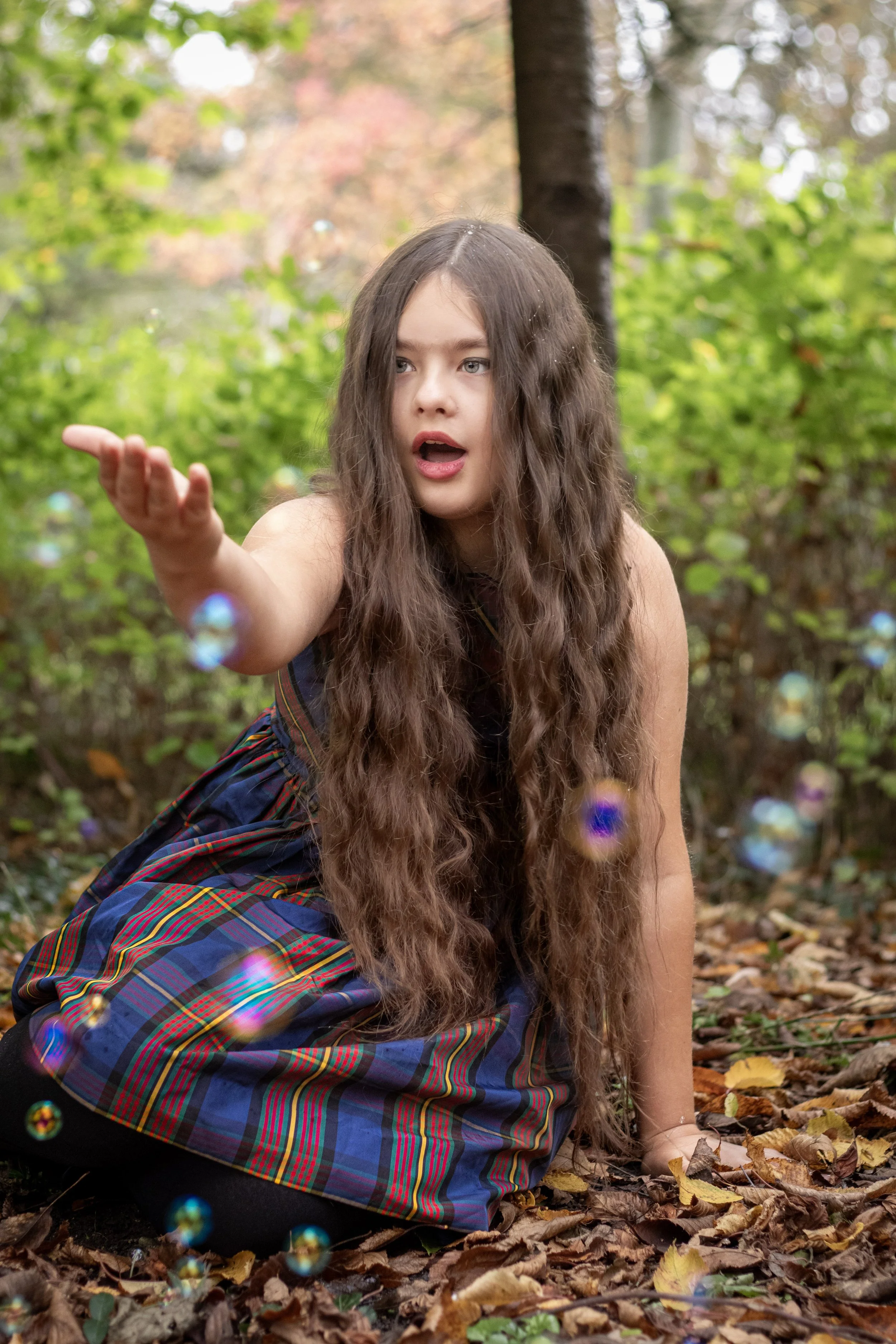 A young girl with long, curly brown hair, wearing a plaid dress, is sitting on the ground in a forest, reaching out and appearing to blow or release bubbles. The background is filled with trees and greenery.