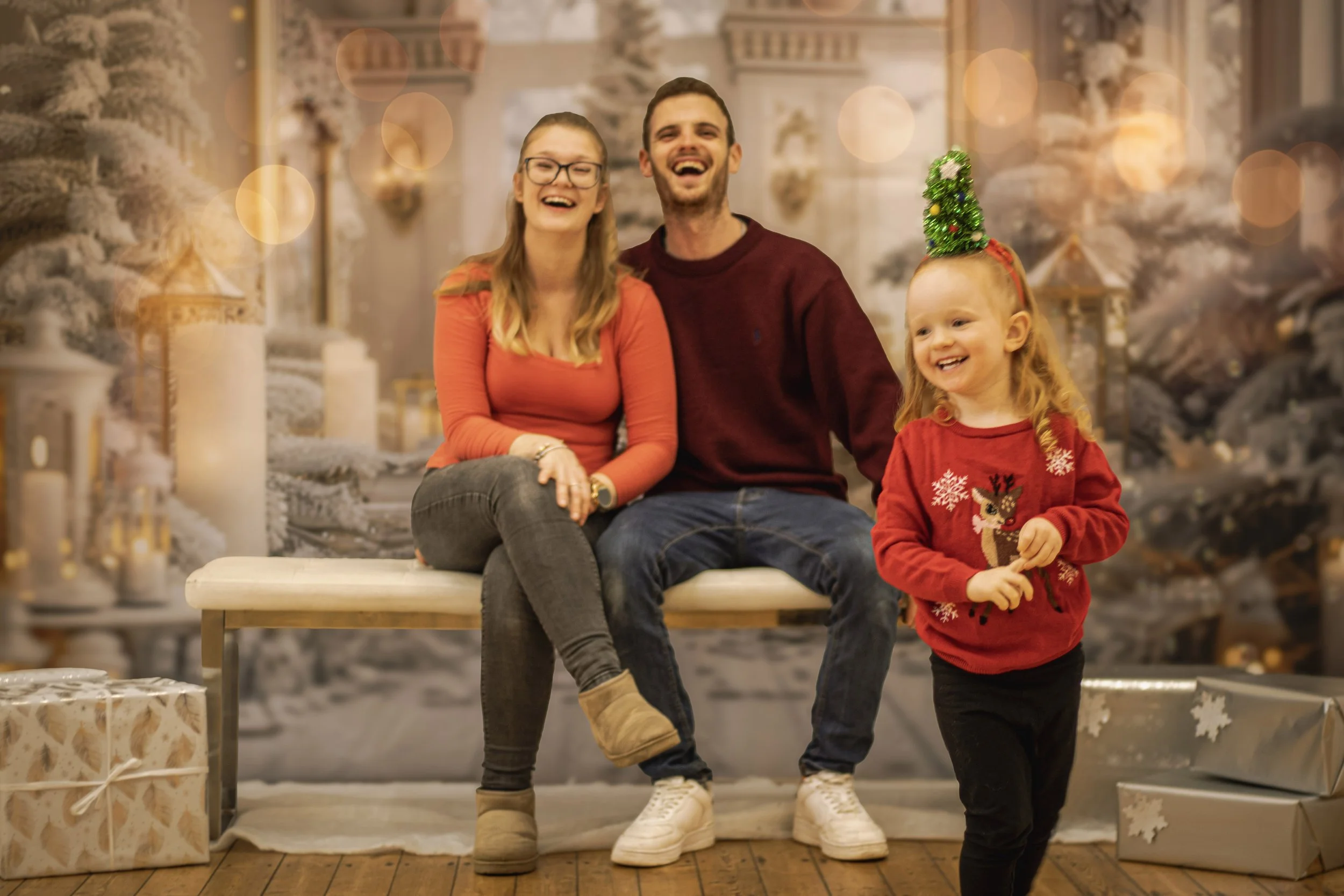 A family of three, including a woman, a man, and a young girl, sitting on a bench in a festive Christmas setting. They are all smiling and dressed in holiday attire, with wrapped presents around them and a decorated Christmas tree in the background.