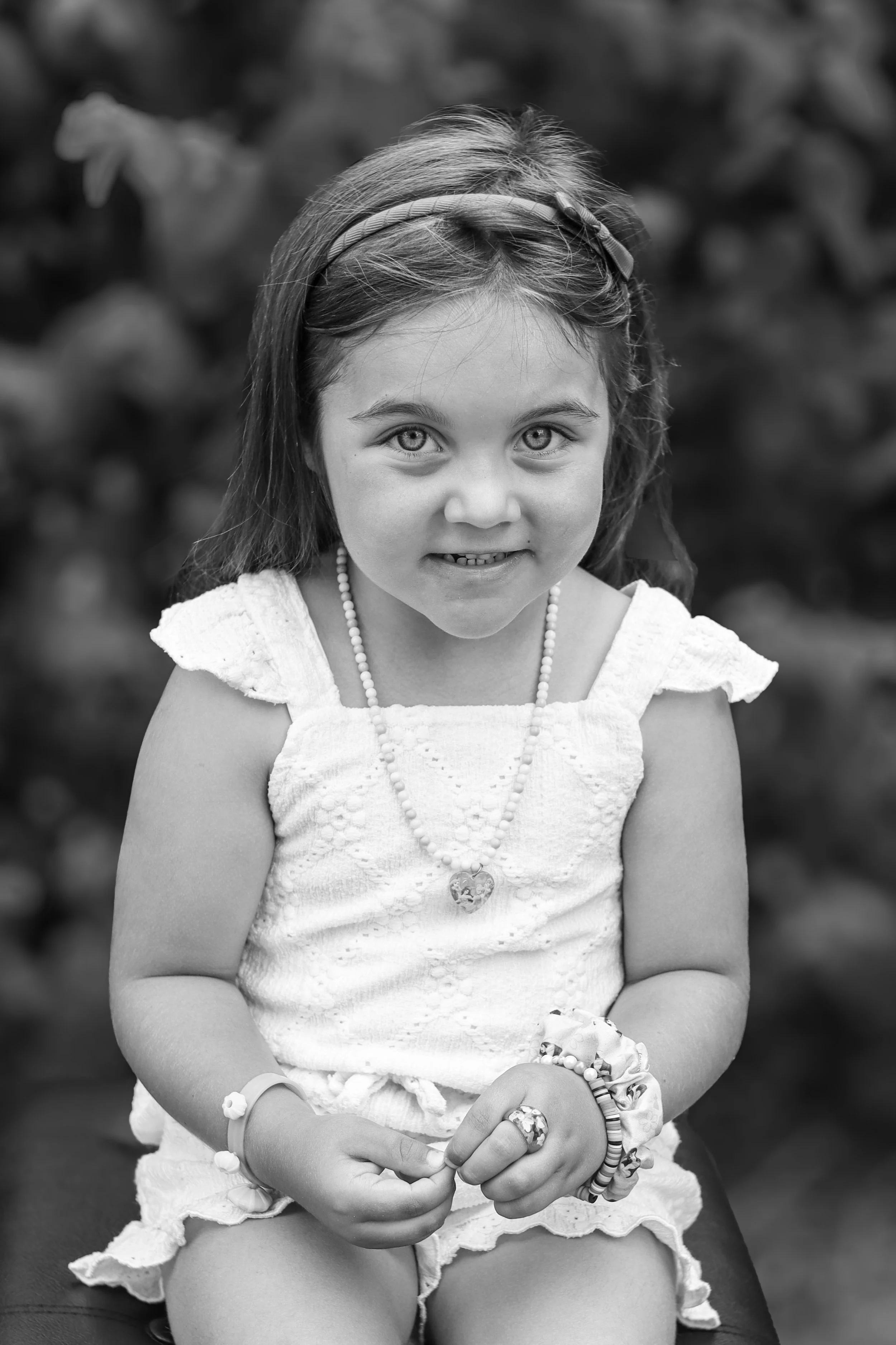 Black and white photo of a young girl with light-colored eyes, wearing a sleeveless dress with ruffled sleeves, a headband, and jewelry including a necklace, bracelets, and a large ring, sitting outdoors with blurred foliage in the background.