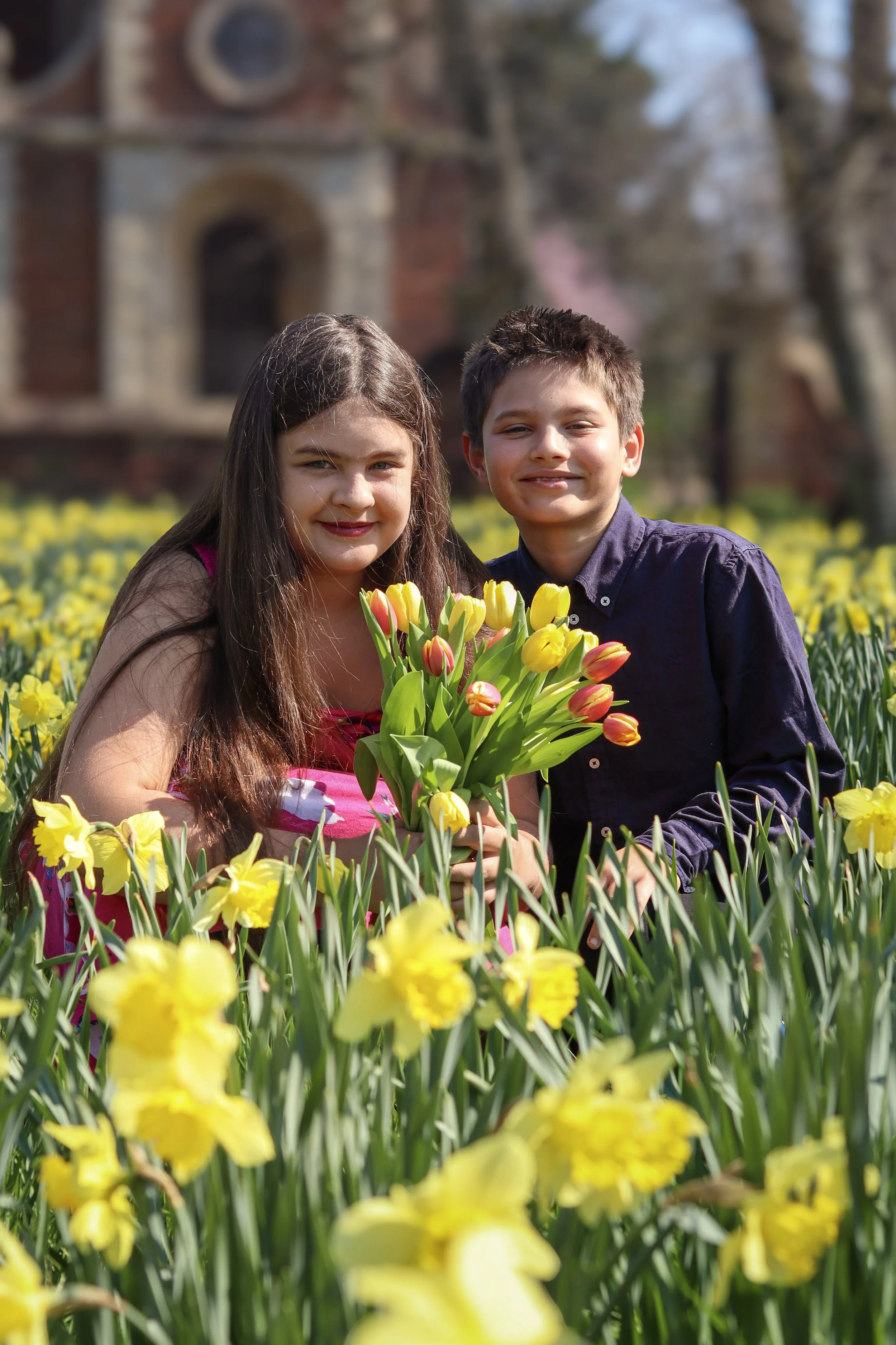 A boy and girl smiling in a yellow daffodil field holding a bouquet of tulips with a church in the background.