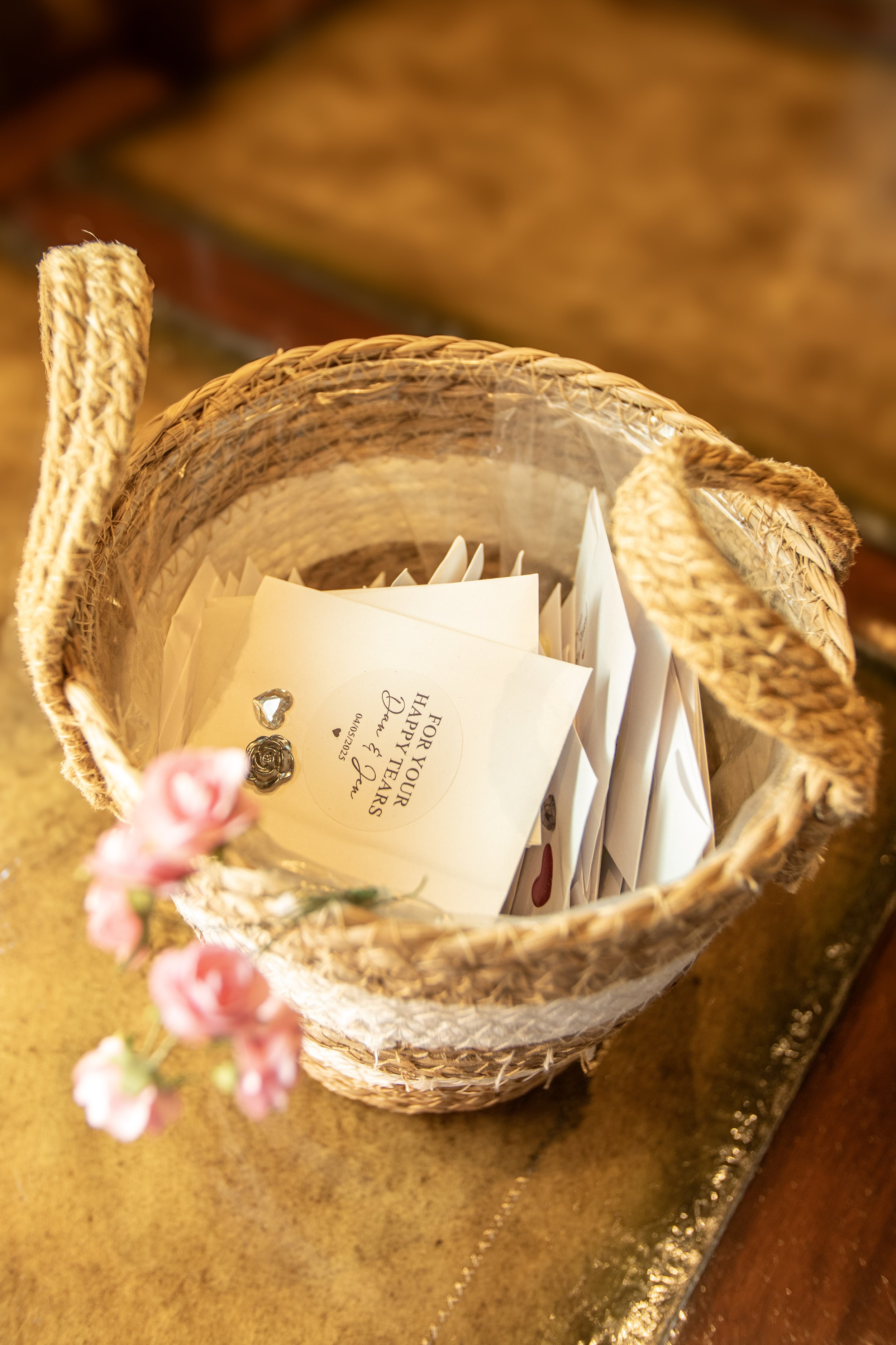 A wicker basket filled with greeting cards, some with visible messages, and decorative jewelry, placed on a shiny surface with a blurred floral decoration in the foreground.