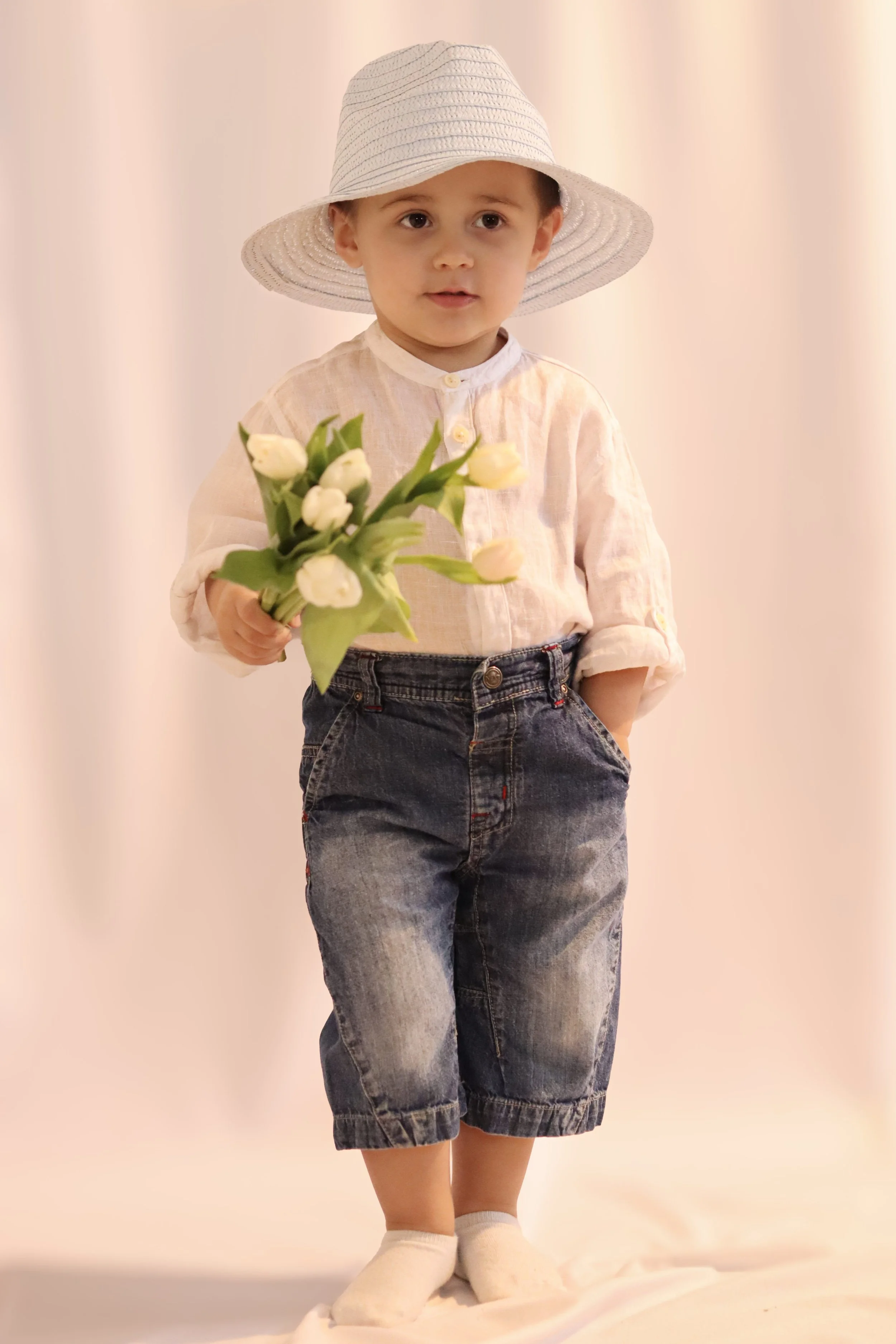 A young boy wearing a white sun hat, a cream-colored long-sleeve shirt, and denim shorts, holding a bouquet of white tulips, standing against a plain light background.