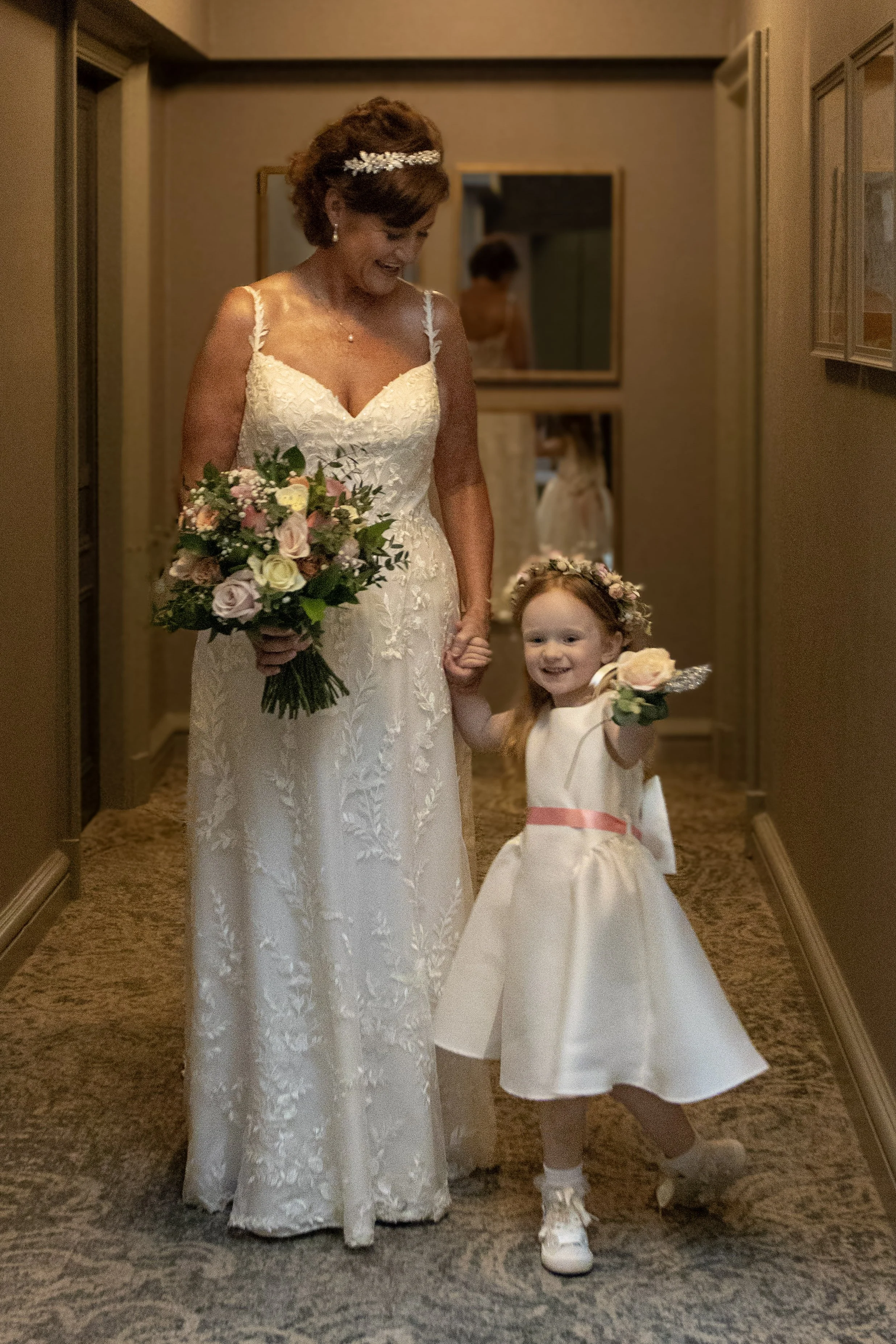 A bride and a young girl holding hands and smiling in a hallway, dressed in wedding attire. The bride holds a bouquet of flowers, and the young girl is wearing a white dress with a pink sash and a floral crown.