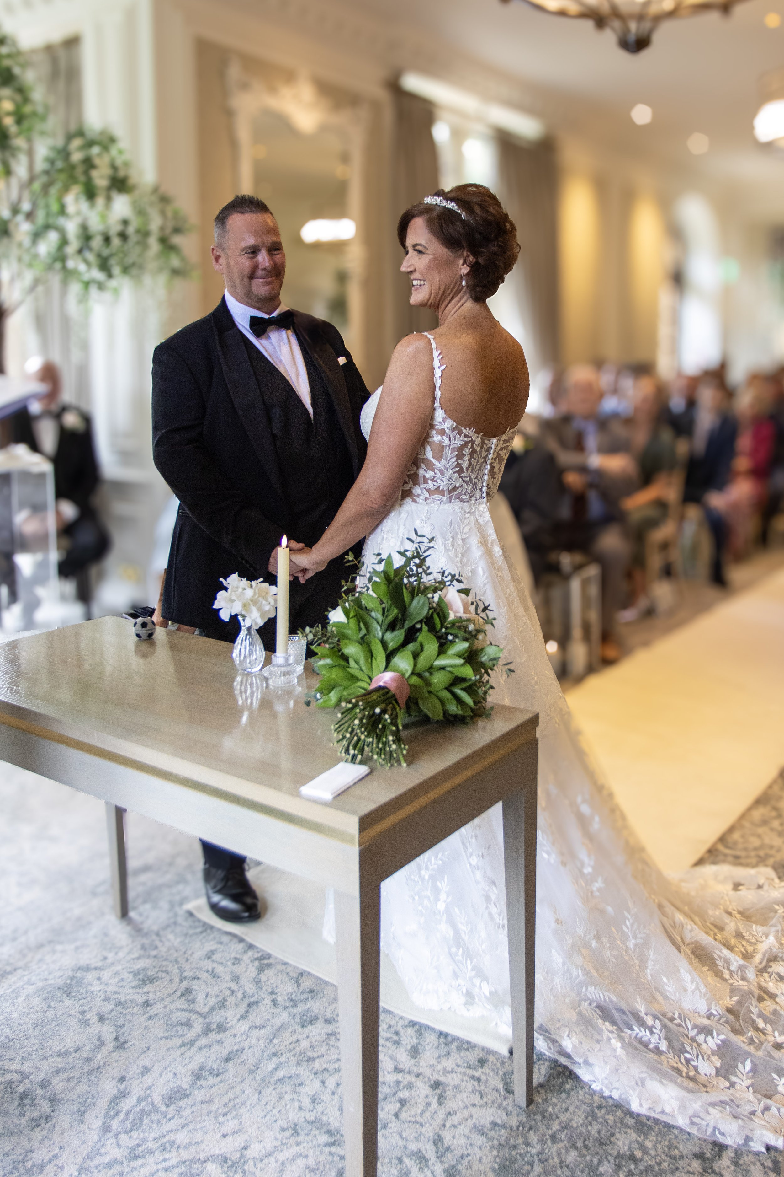 Bride and groom exchanging vows during their wedding ceremony in an elegant room with seated guests in the background.