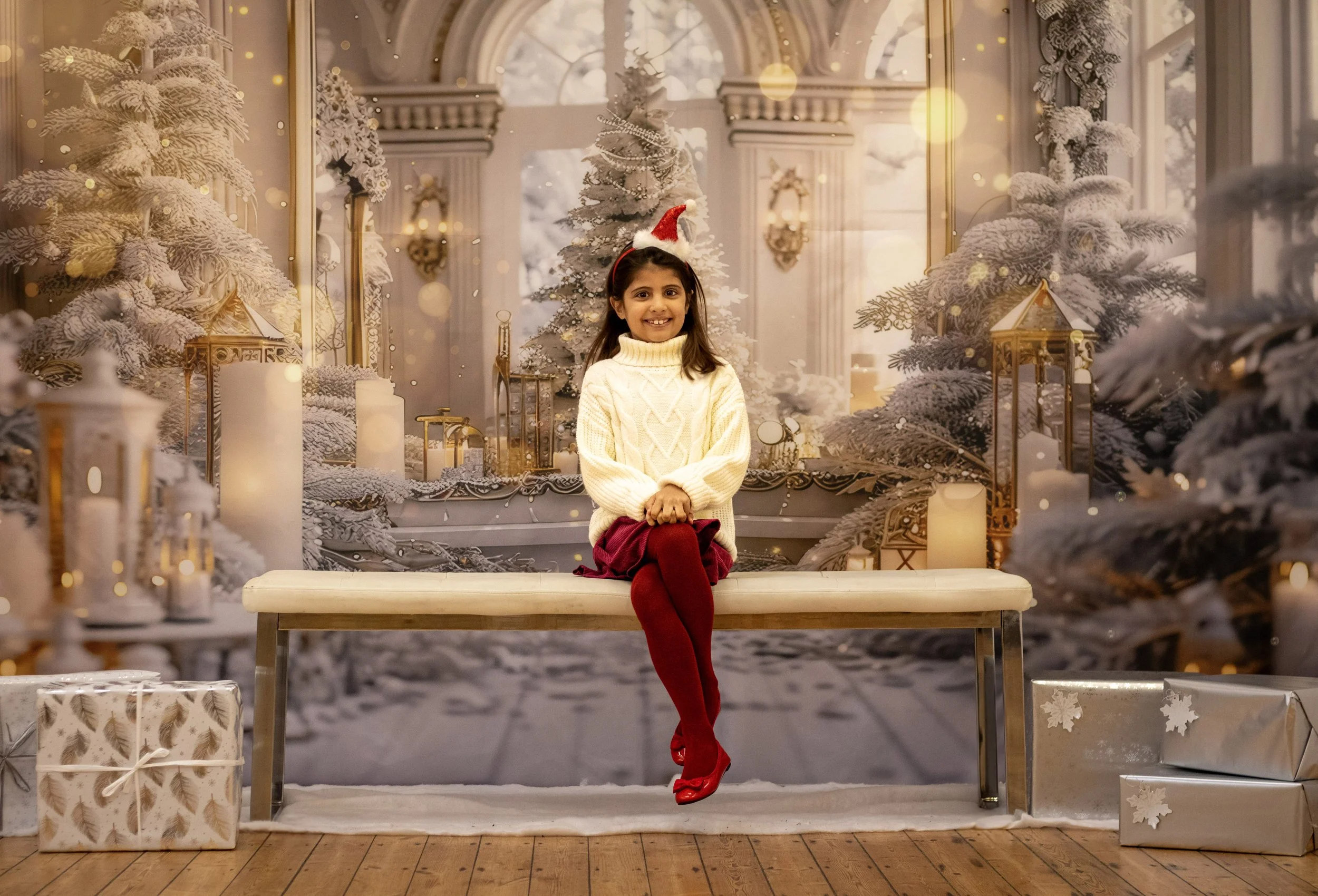 A young girl with dark hair, wearing a red Santa hat, cream-colored sweater, red skirt, and red tights, sitting on a white bench with a holiday-themed background featuring snow-covered trees, white candles, lanterns, and wrapped presents.
