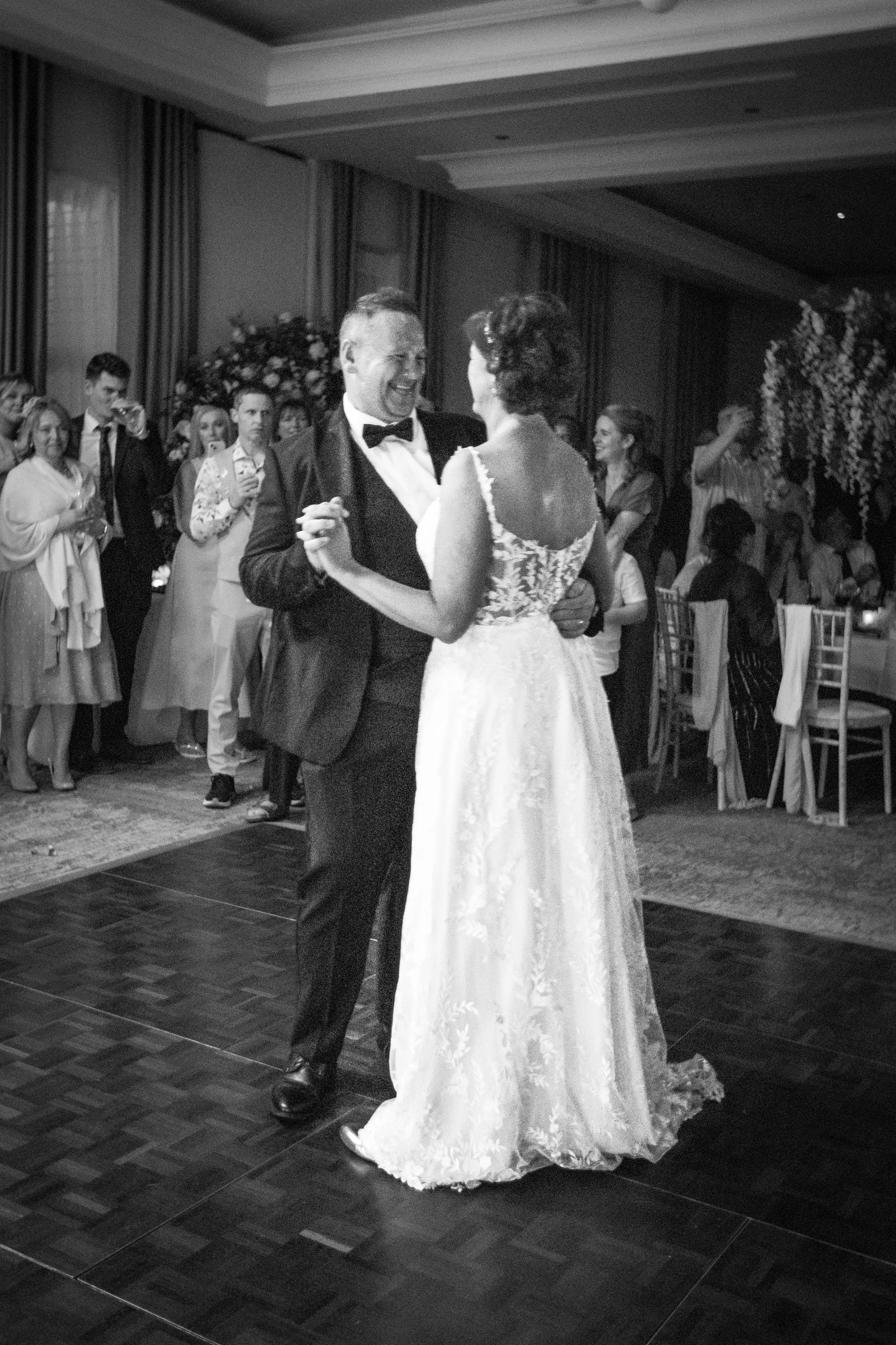 A black and white photo of a bride and groom dancing at their wedding reception, surrounded by guests.