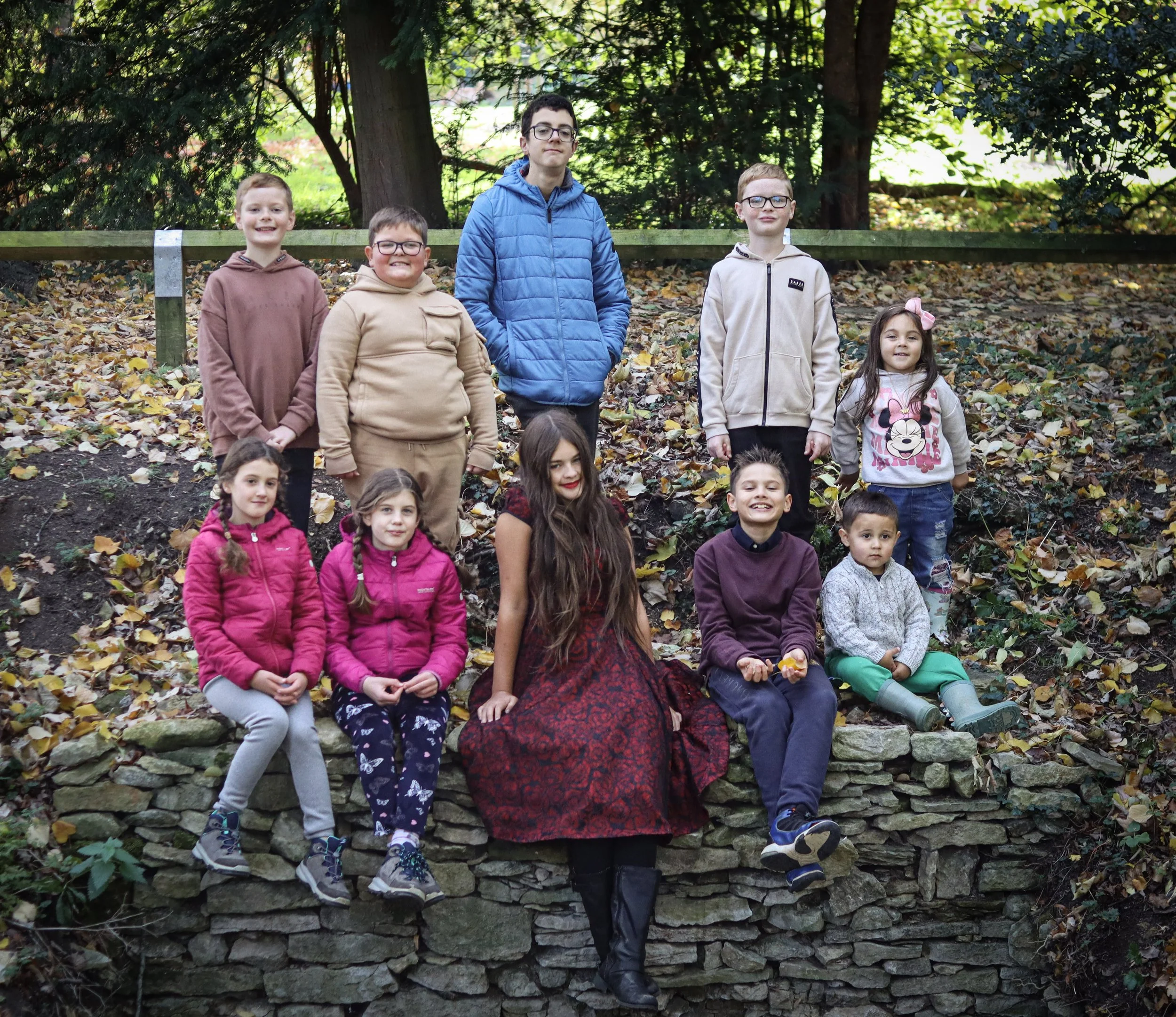 A group of ten children outdoors in a wooded area, sitting and standing on a stone wall and hillside, with trees and fallen leaves in the background.